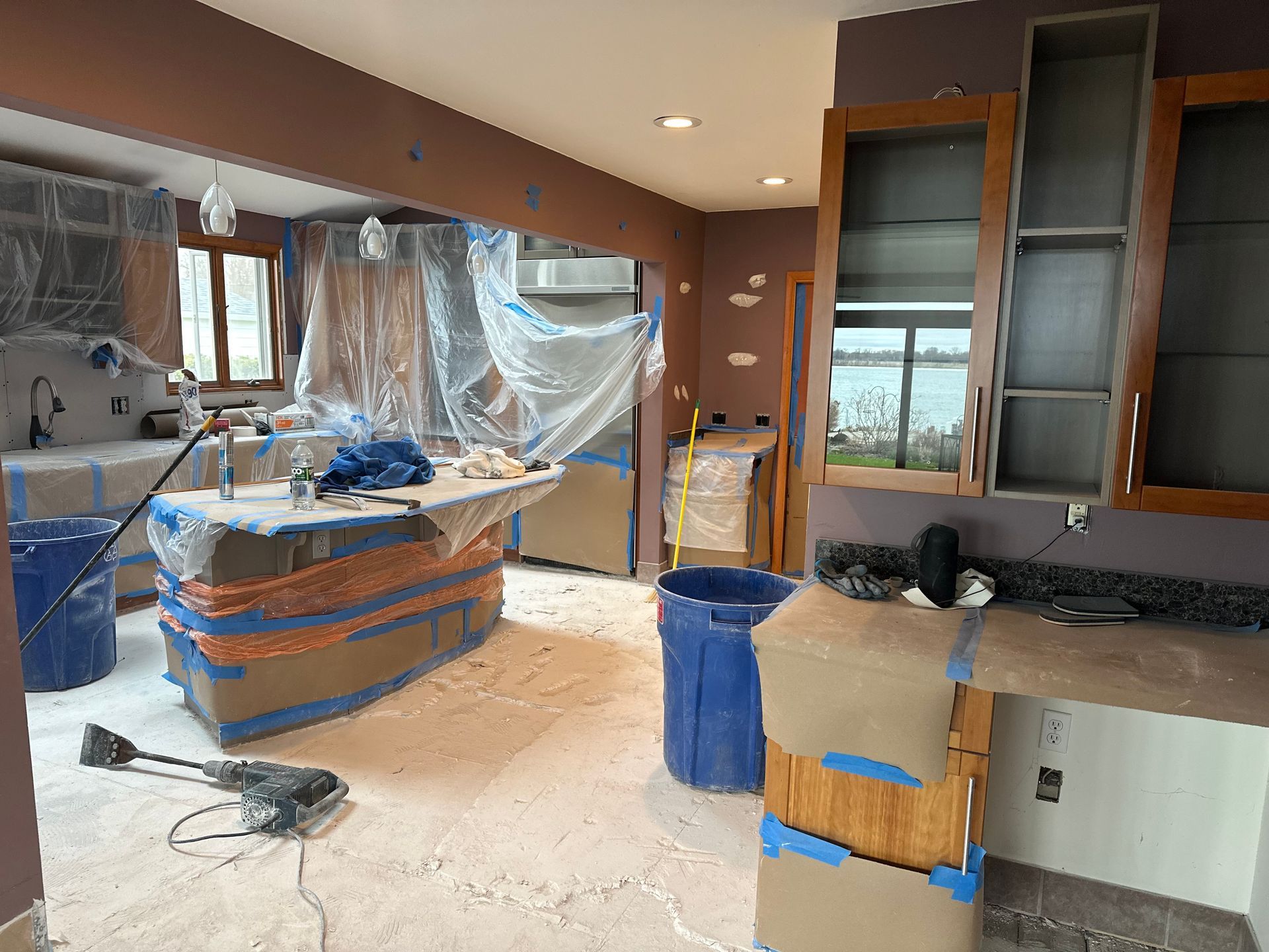 Kitchen under construction with cabinets and island covered in plastic and masking tape, with debris on the floor.