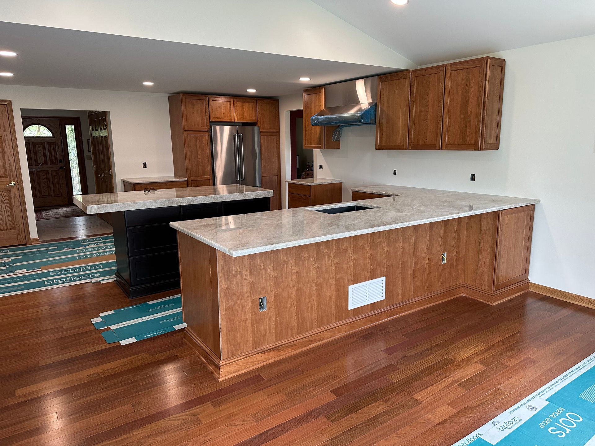 A kitchen with wooden cabinets, light stone countertops, a central island, stainless steel fridge, and hardwood floors.