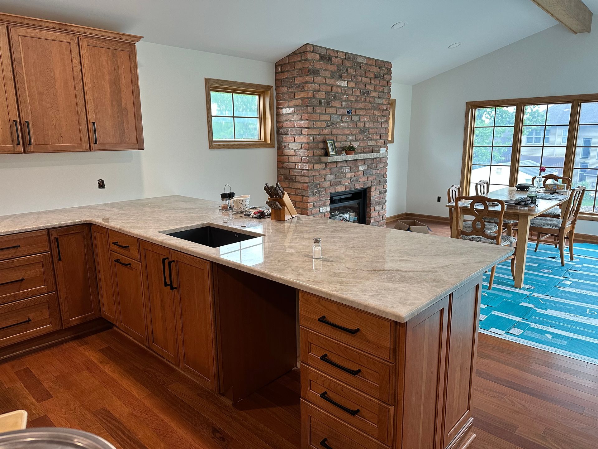 A kitchen with oak cabinets, a light stone countertop with an inset sink, a brick fireplace, and a dining area beyond.