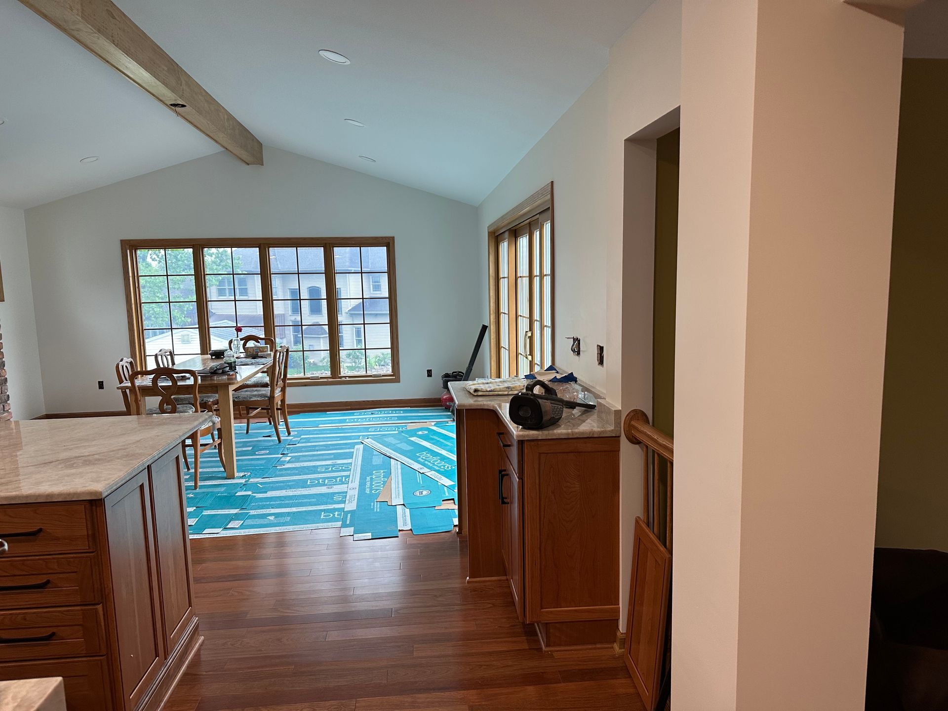 A kitchen island with wood cabinets and granite countertops overlooks a dining area with blue floor protection film.