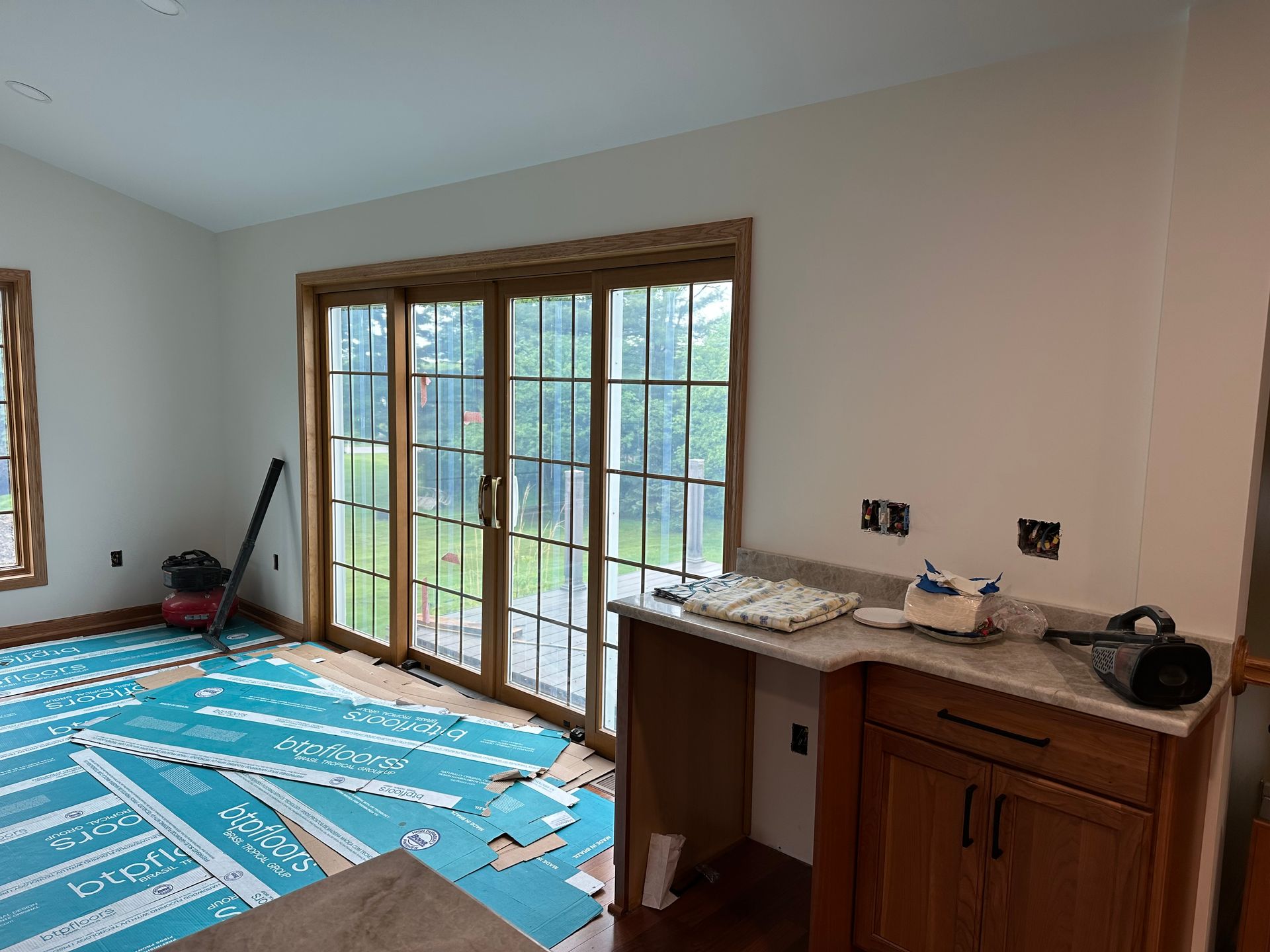 A bright room under renovation with wood-trimmed sliding glass doors, wooden cabinetry, and blue flooring protective film.