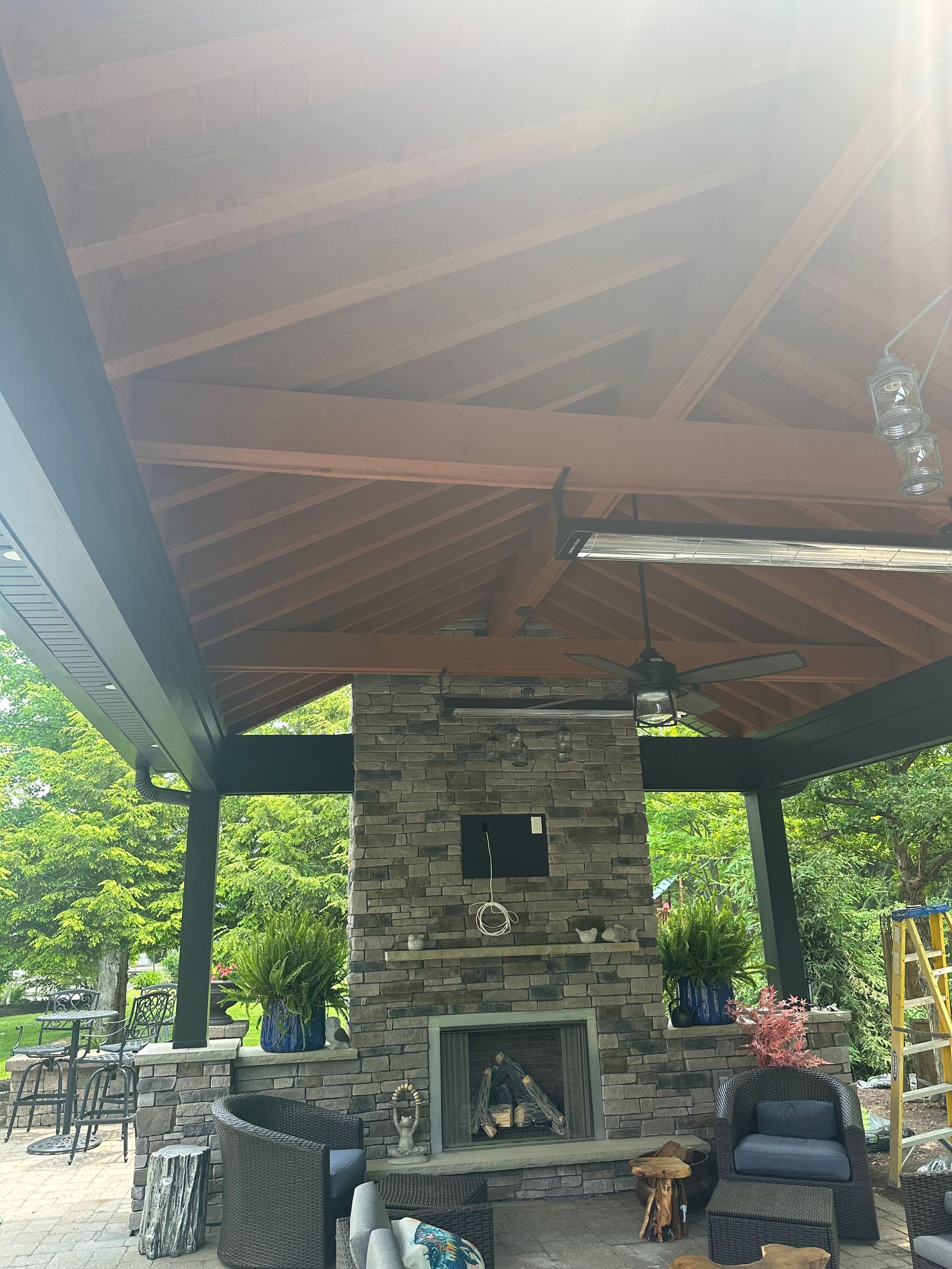 A stone fireplace under a wooden pavilion, featuring outdoor furniture and greenery.