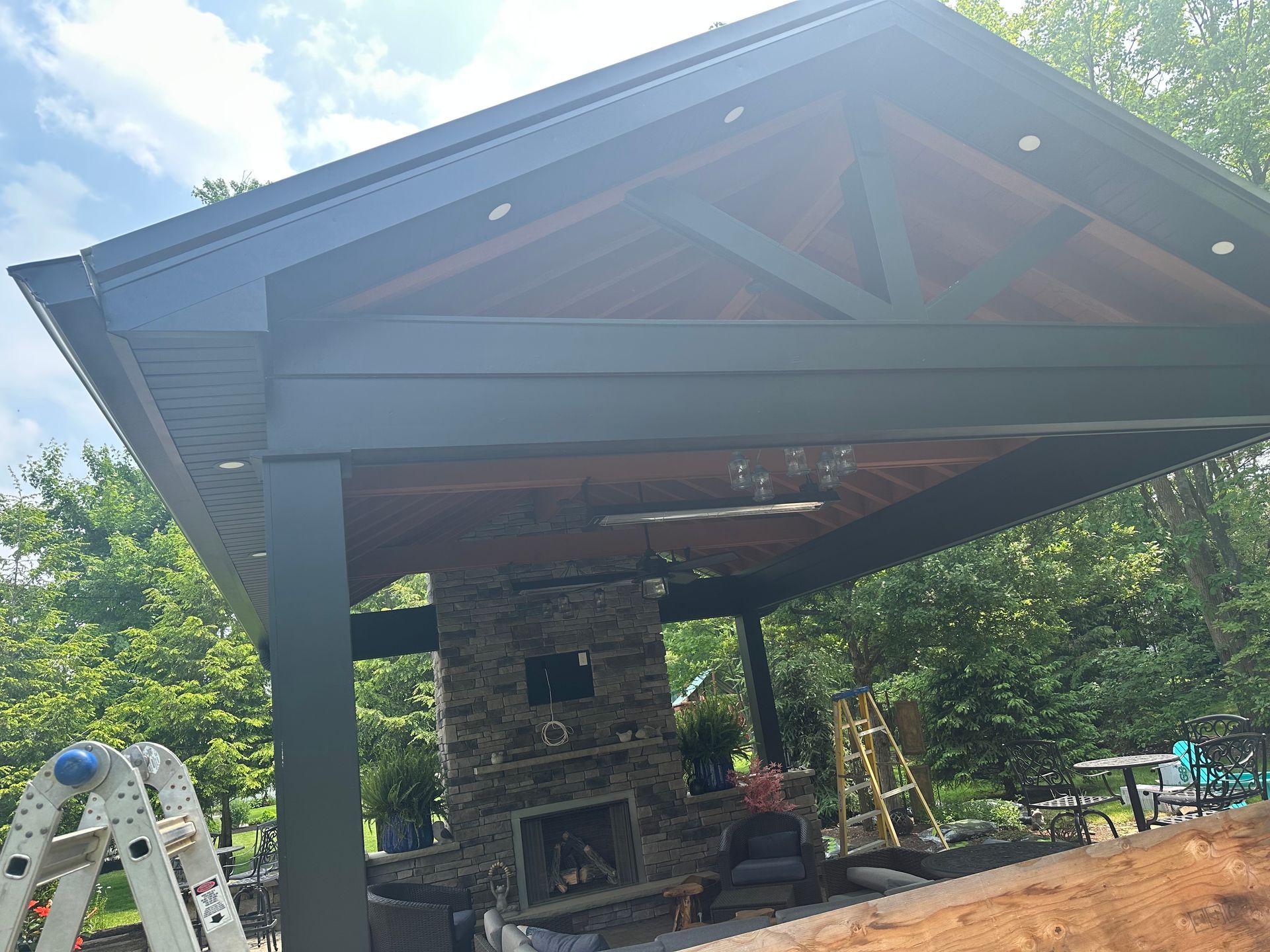 An outdoor pavilion with a dark frame, stone fireplace, and wood-paneled ceiling set in a lush, green backyard.
