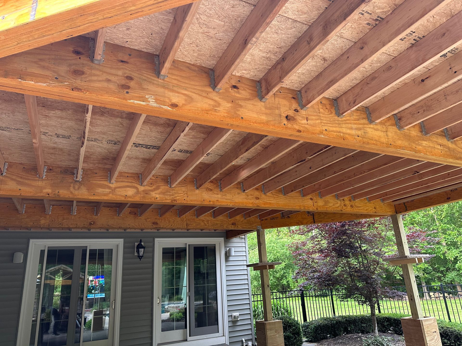 An outdoor patio covered by an exposed wooden roof structure with joists, looking toward a gray house with glass doors.