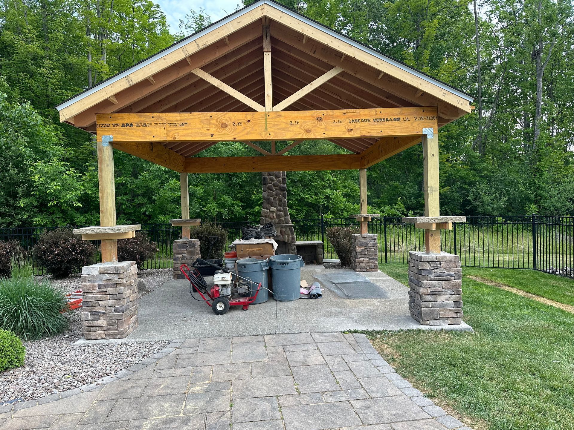A wooden open-air pavilion with a metal roof stands over a stone patio featuring stone columns and a central grill station.