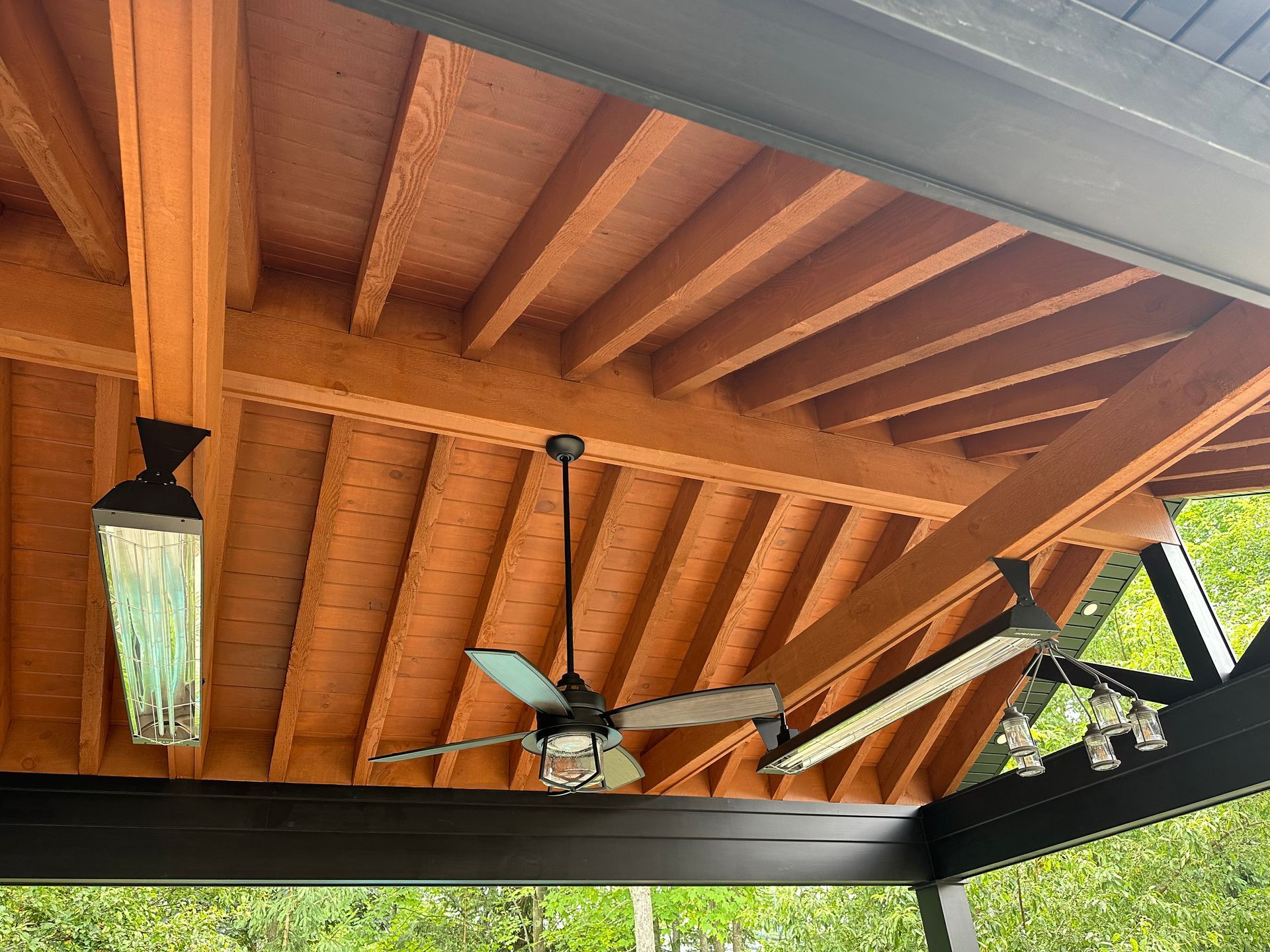 An outdoor wooden ceiling with exposed beams, a ceiling fan, two linear heaters, and a small cluster of light fixtures.