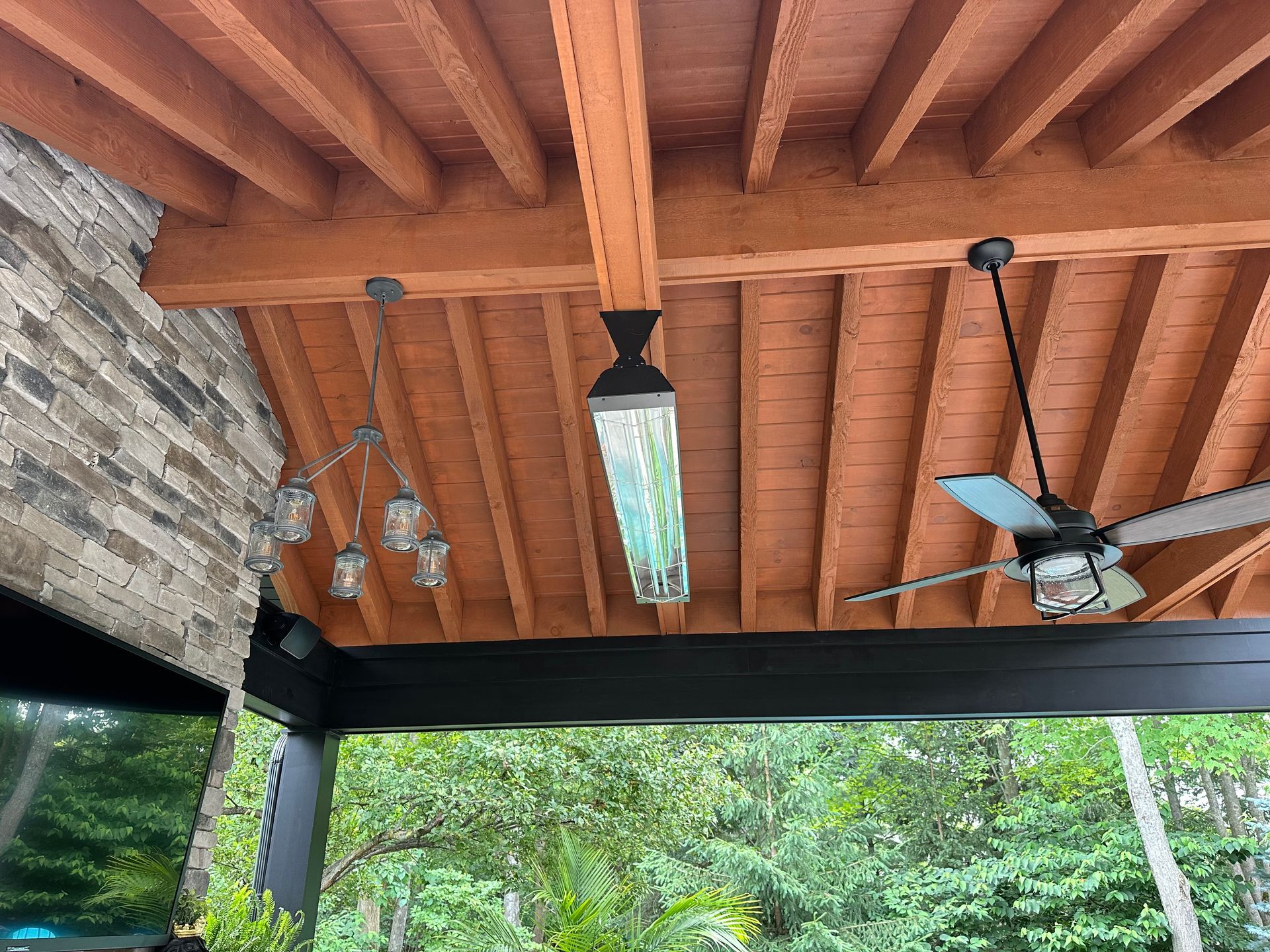 A wooden ceiling with an outdoor heater, a hanging pendant light fixture, and a ceiling fan over a patio with greenery.