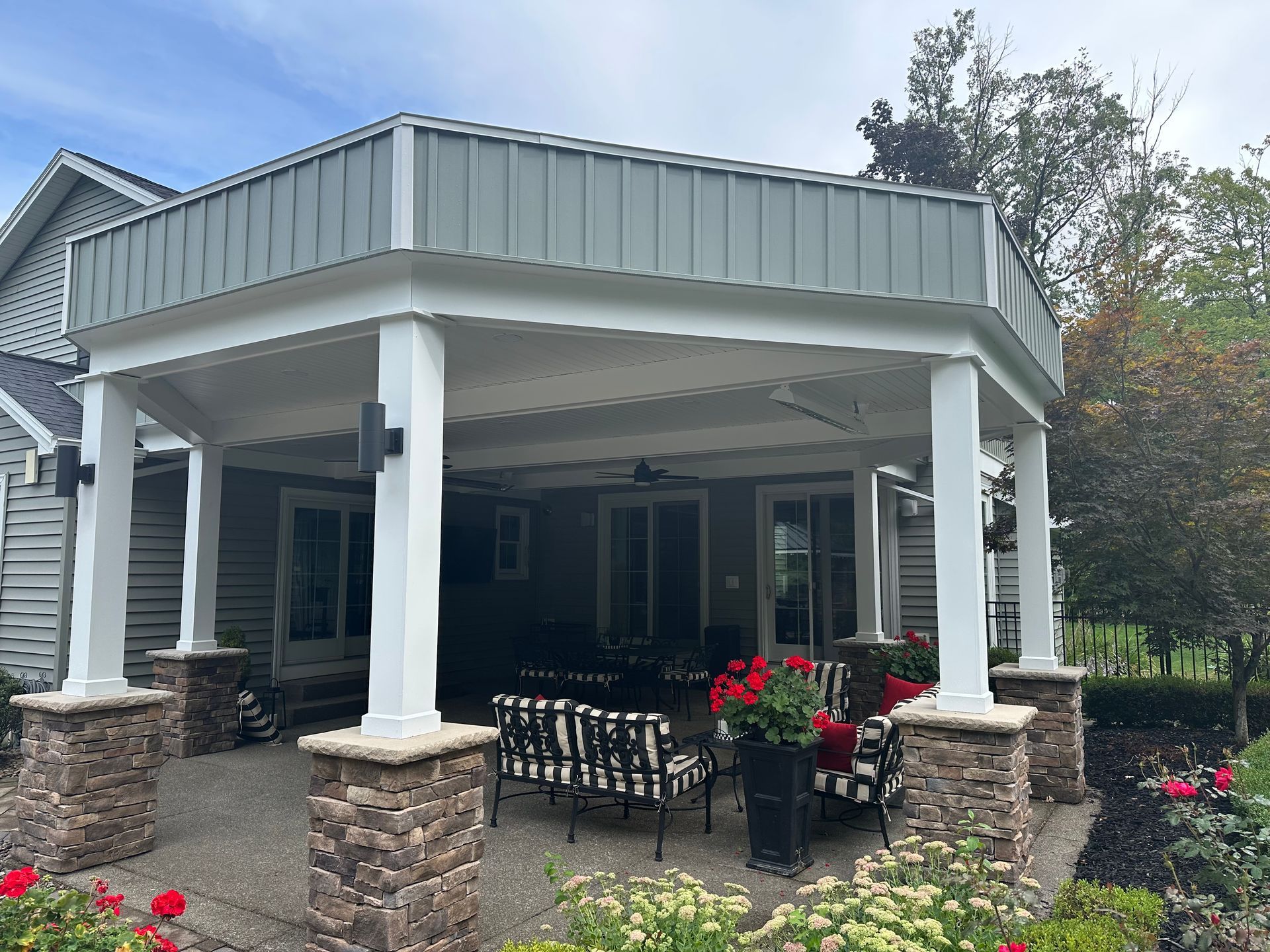 A covered patio with a gray roof and white columns over stone bases, featuring outdoor furniture and flower pots.