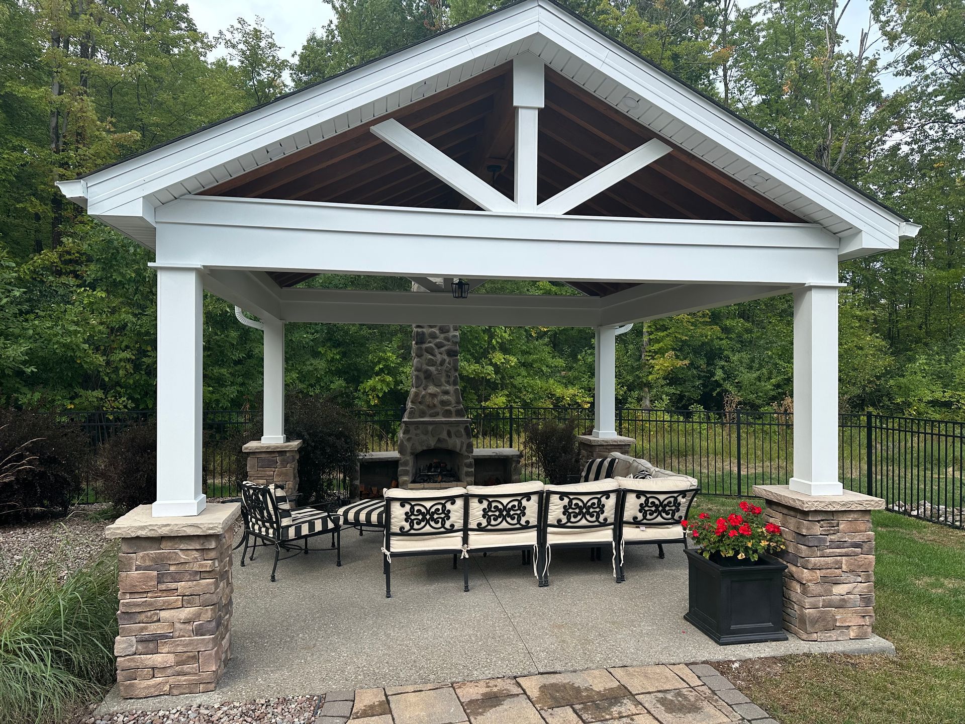 A covered outdoor pavilion with a stone fireplace, patio furniture, and a red flower planter on a stone patio.