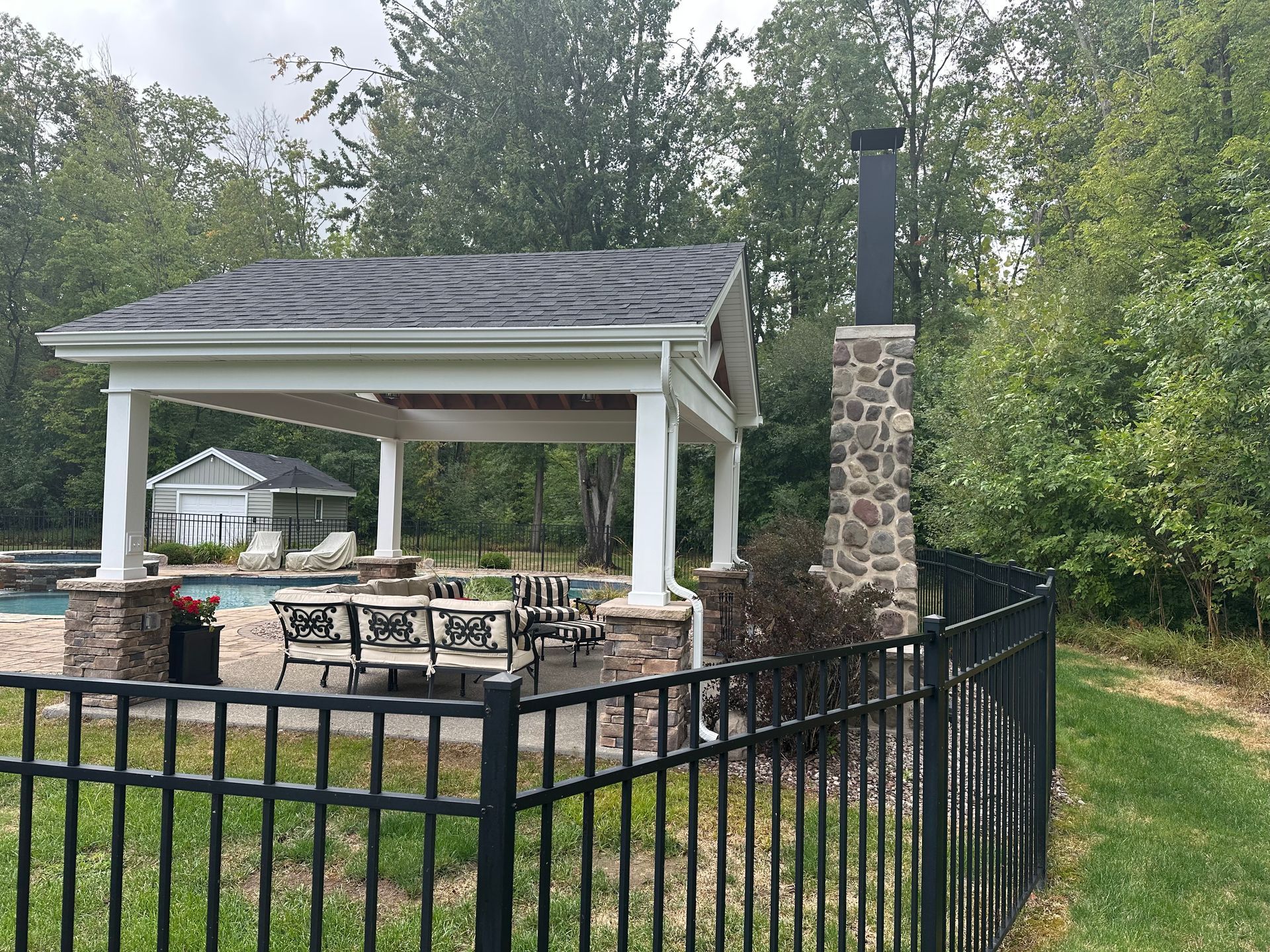 A stone-pillared pavilion with a shingled roof and fireplace stands next to a pool behind a black metal fence.