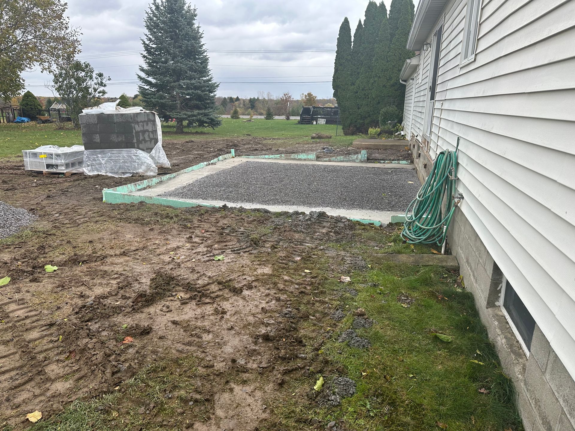 A rectangular gravel base for a home addition, enclosed by green insulation forms, situated next to a white house.