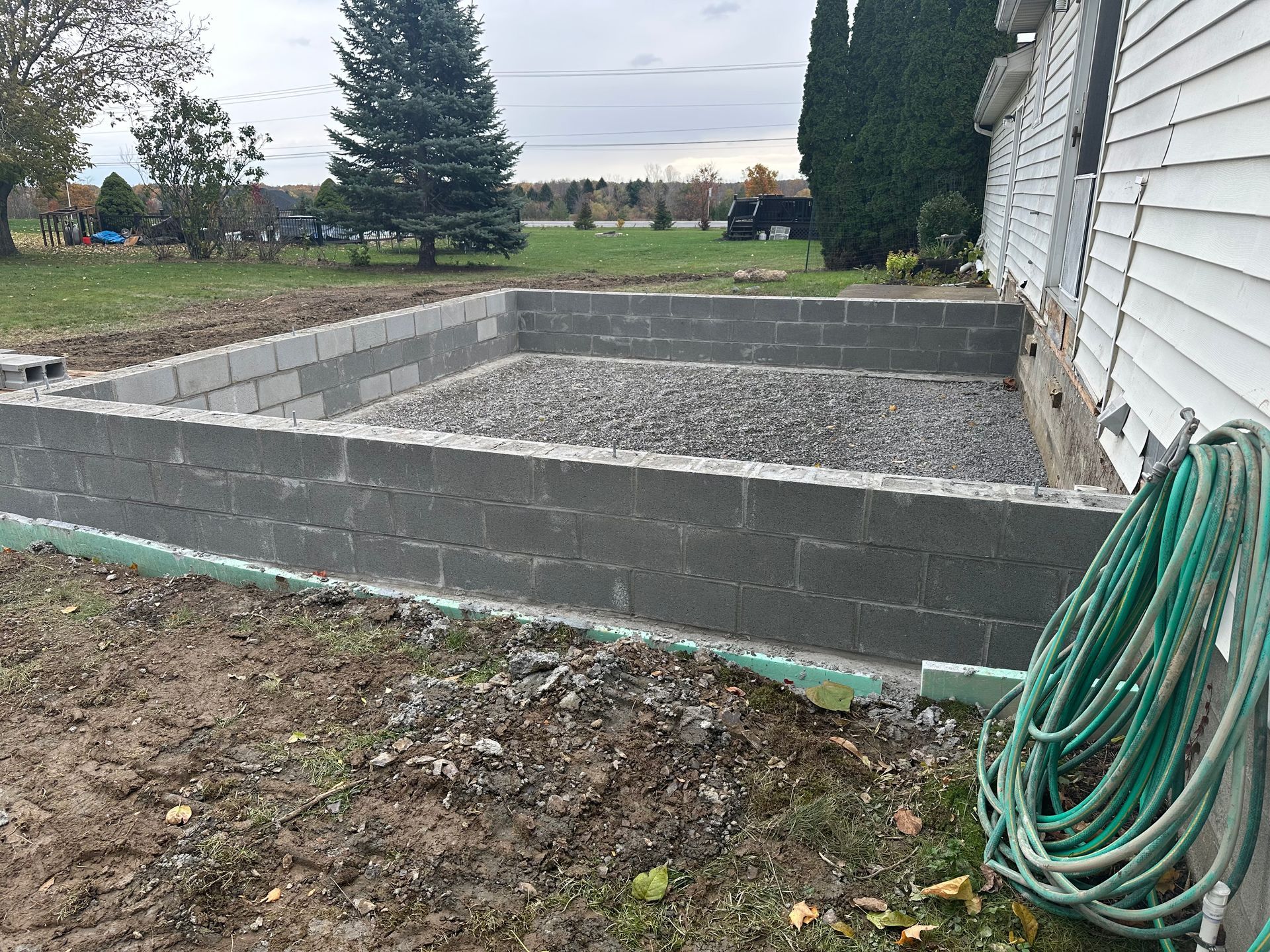 A cinder block foundation for a building addition in a grassy yard, with a gravel interior base and a green garden hose.