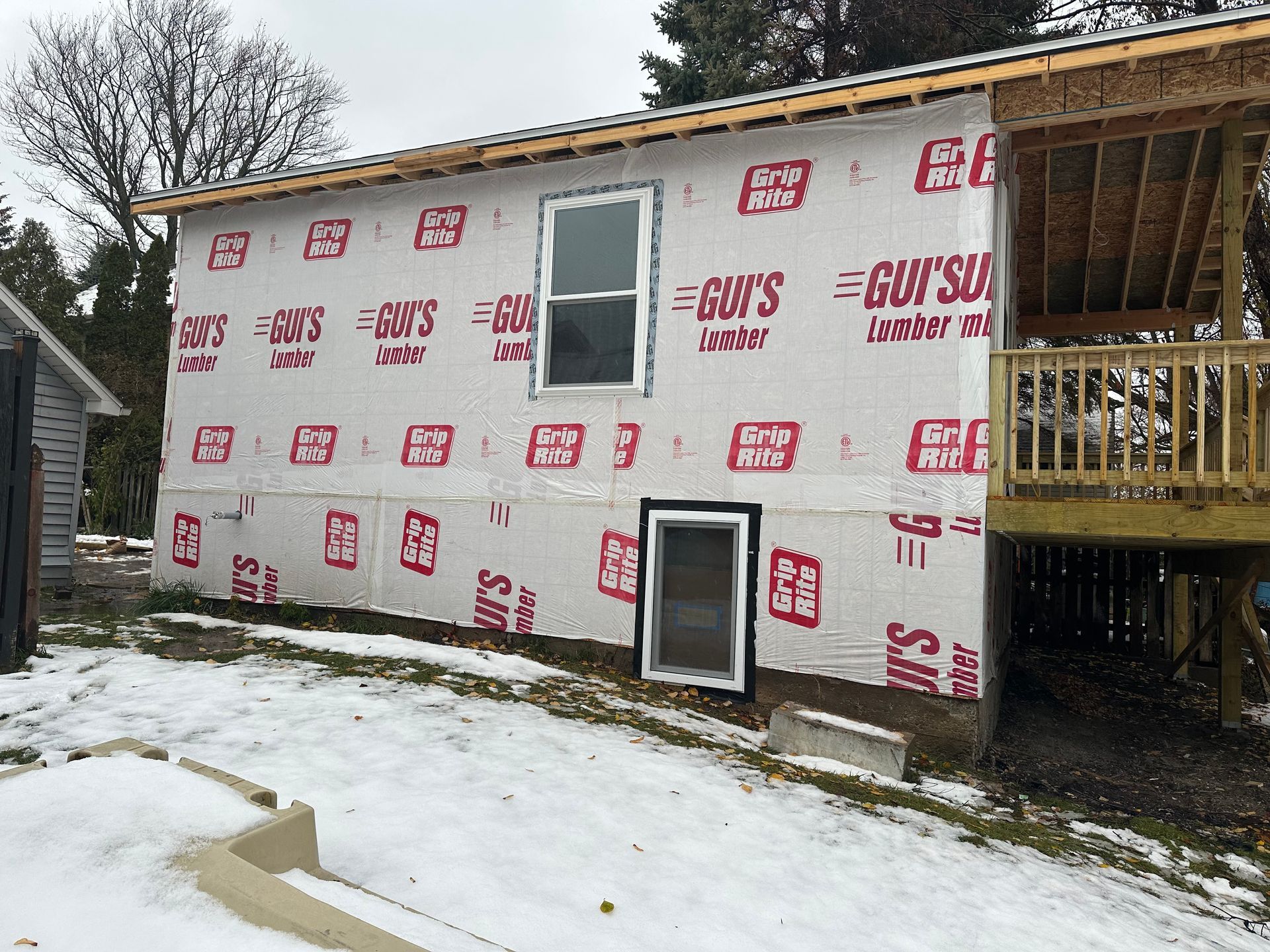 Exterior view of a house under renovation with white vapor barrier siding, two windows, and an adjacent wooden deck.
