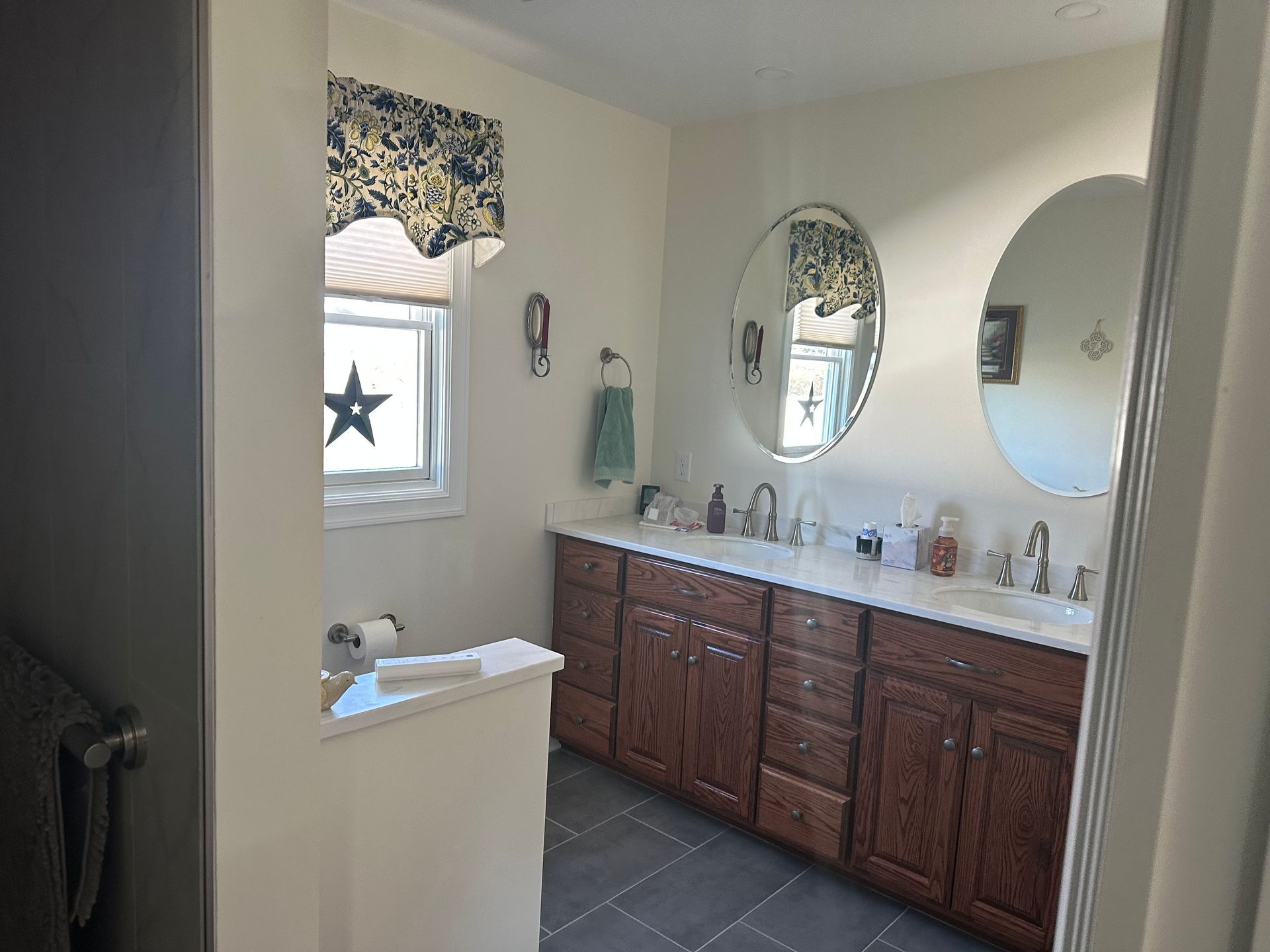 Bathroom with dark wood double vanity, marble-style countertop, two oval mirrors, and a window with a star decoration.