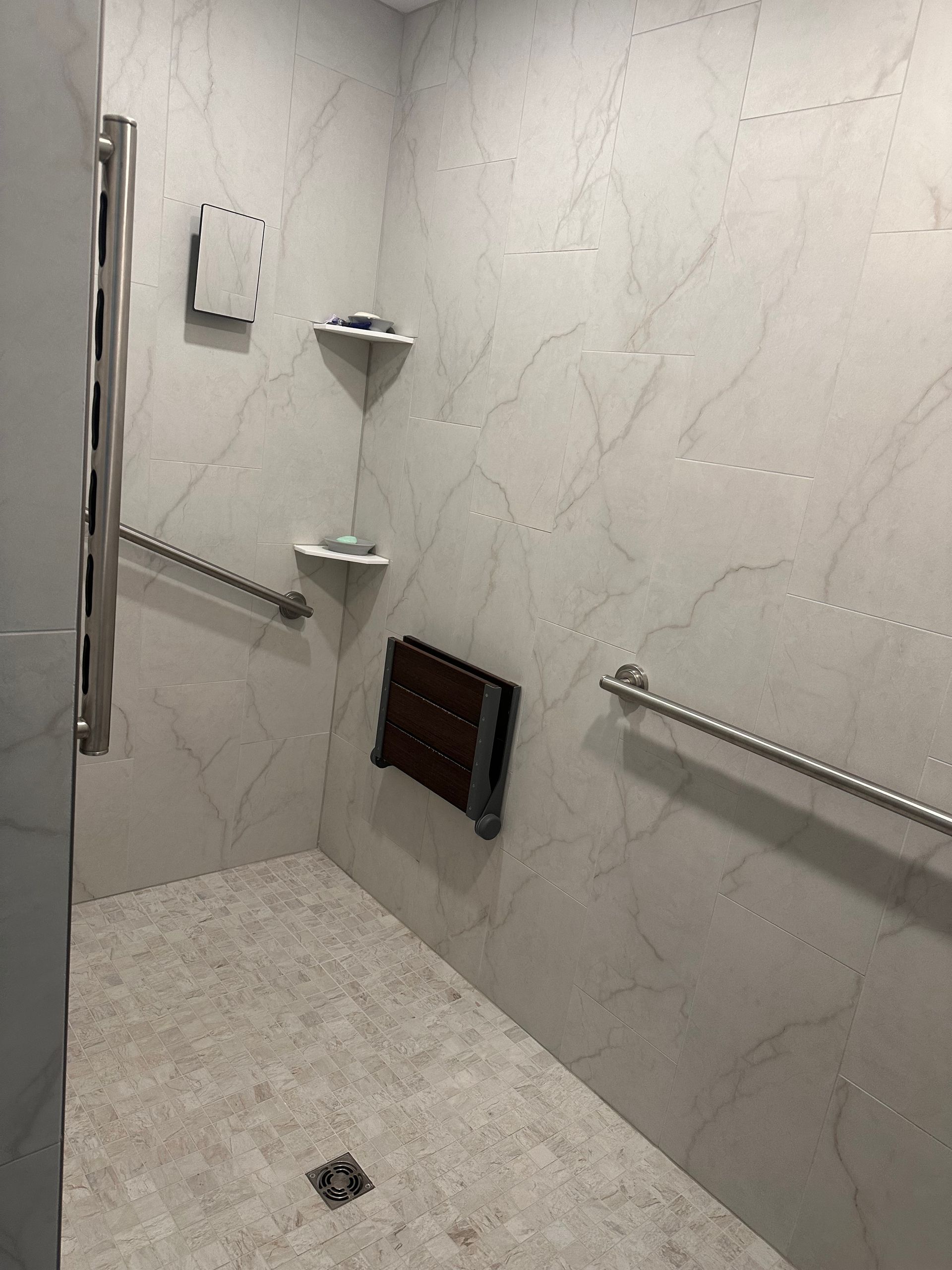 An accessible walk-in shower with light marble-style tiles, a folding wooden seat, two metal grab bars, and corner shelves.
