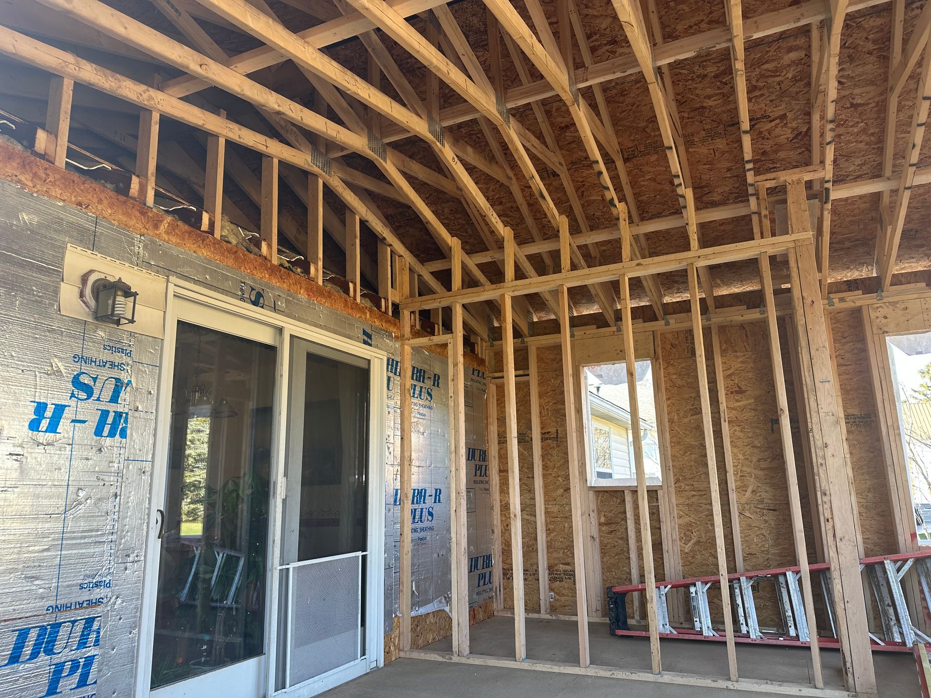 A construction site showing wood framing for a wall and roof structure, with a sliding glass door and exposed insulation.