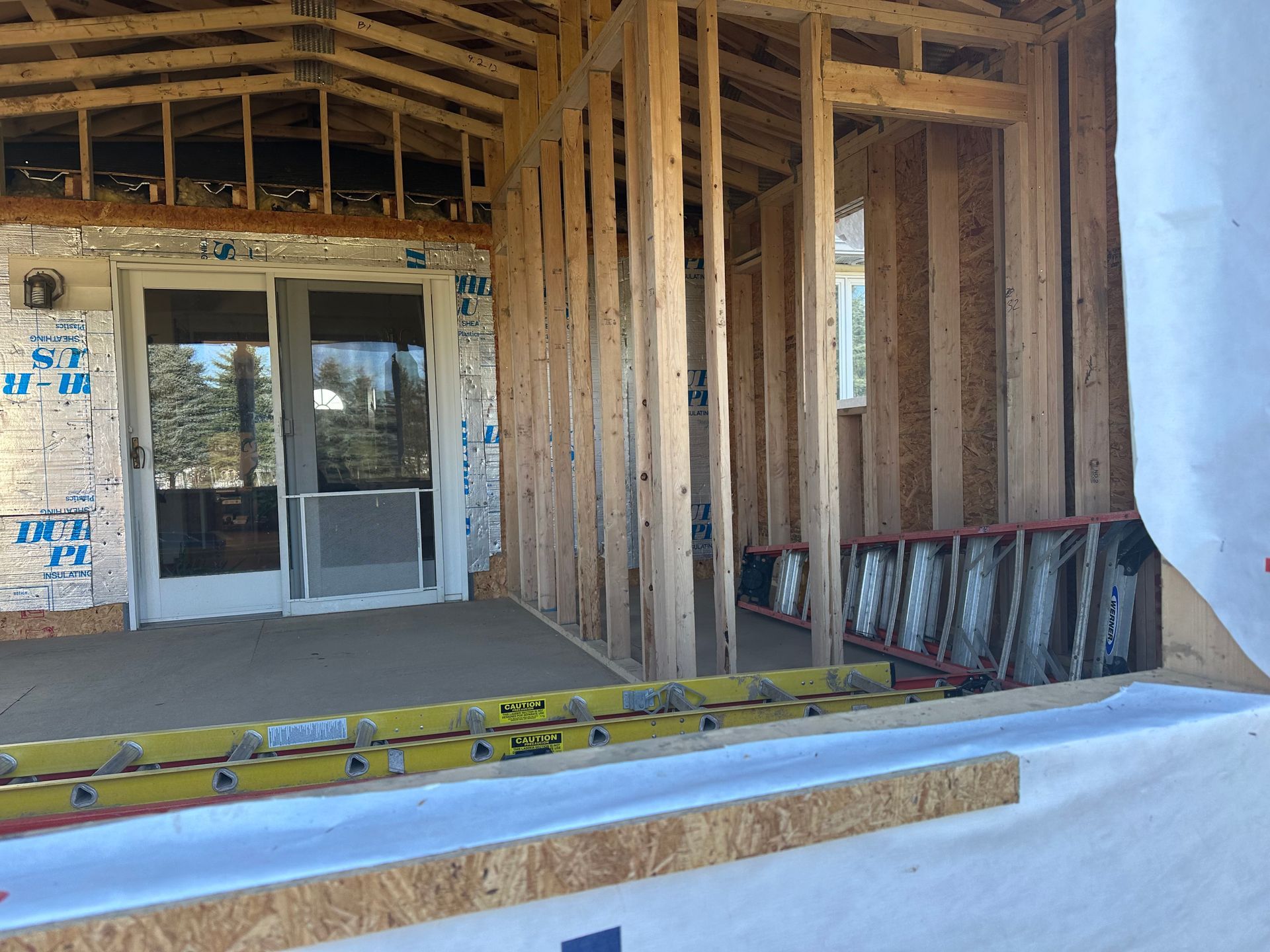 A construction site showing a house frame with exposed wooden studs, a sliding glass door, and yellow ladders.
