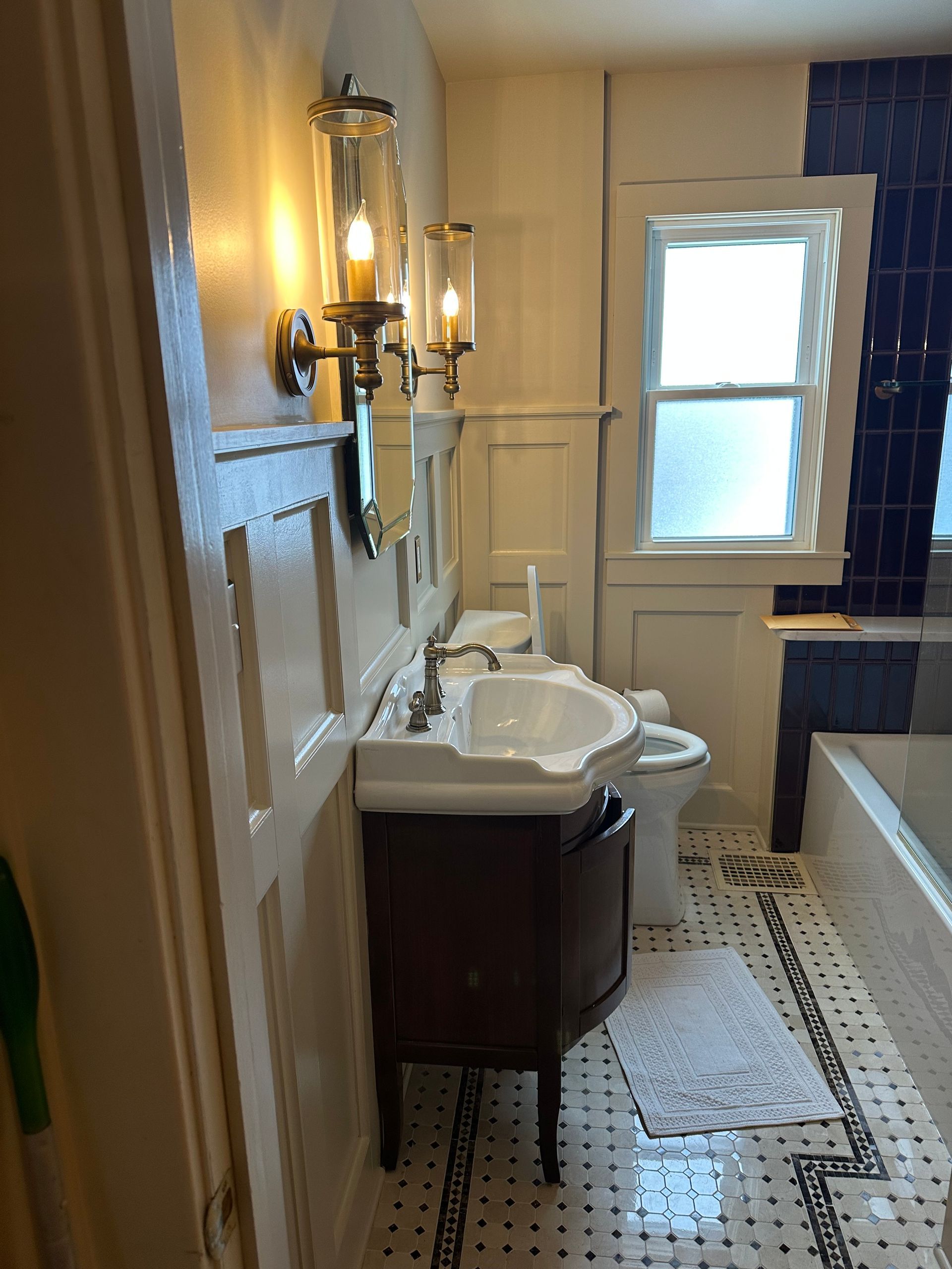 A bathroom featuring a dark wood vanity with a white porcelain sink, wall sconces, patterned floor tile, and a tub.
