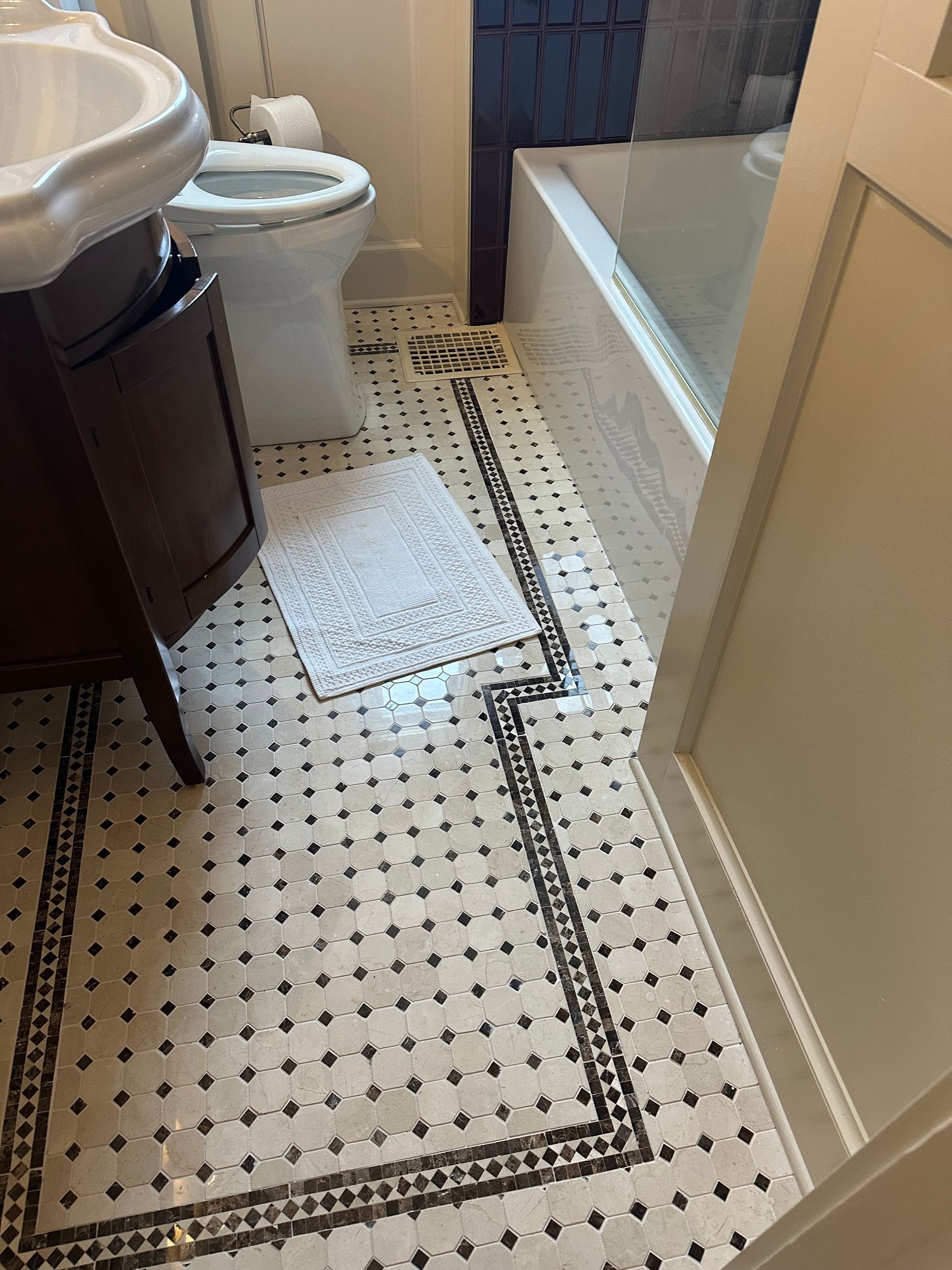 A bathroom floor with black and white basketweave tile, a dark wood vanity, a white toilet, and a light-colored bath mat.