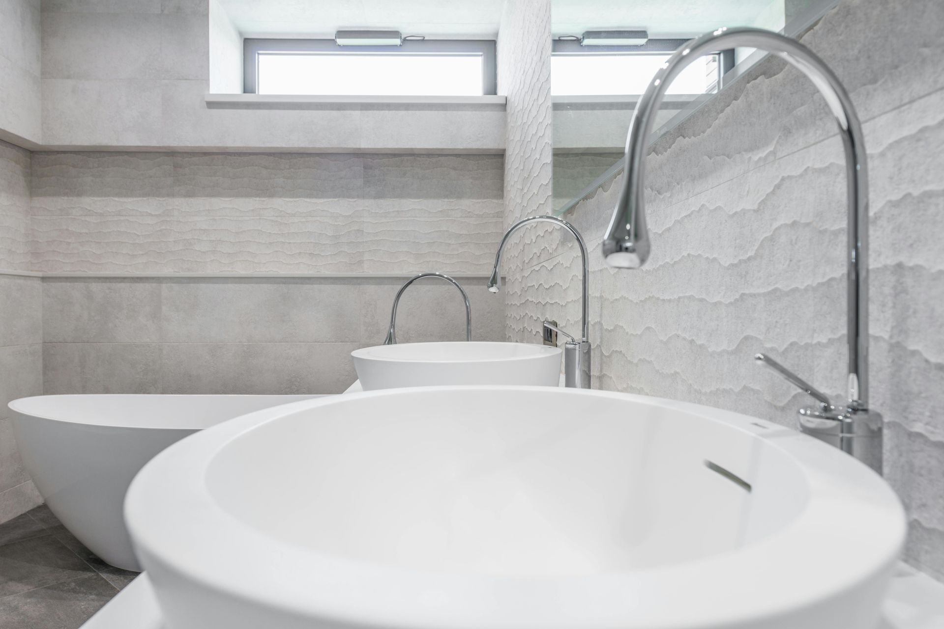 Modern bathroom with dual white vessel sinks, chrome faucets, and textured gray tiled walls, with a freestanding tub nearby.