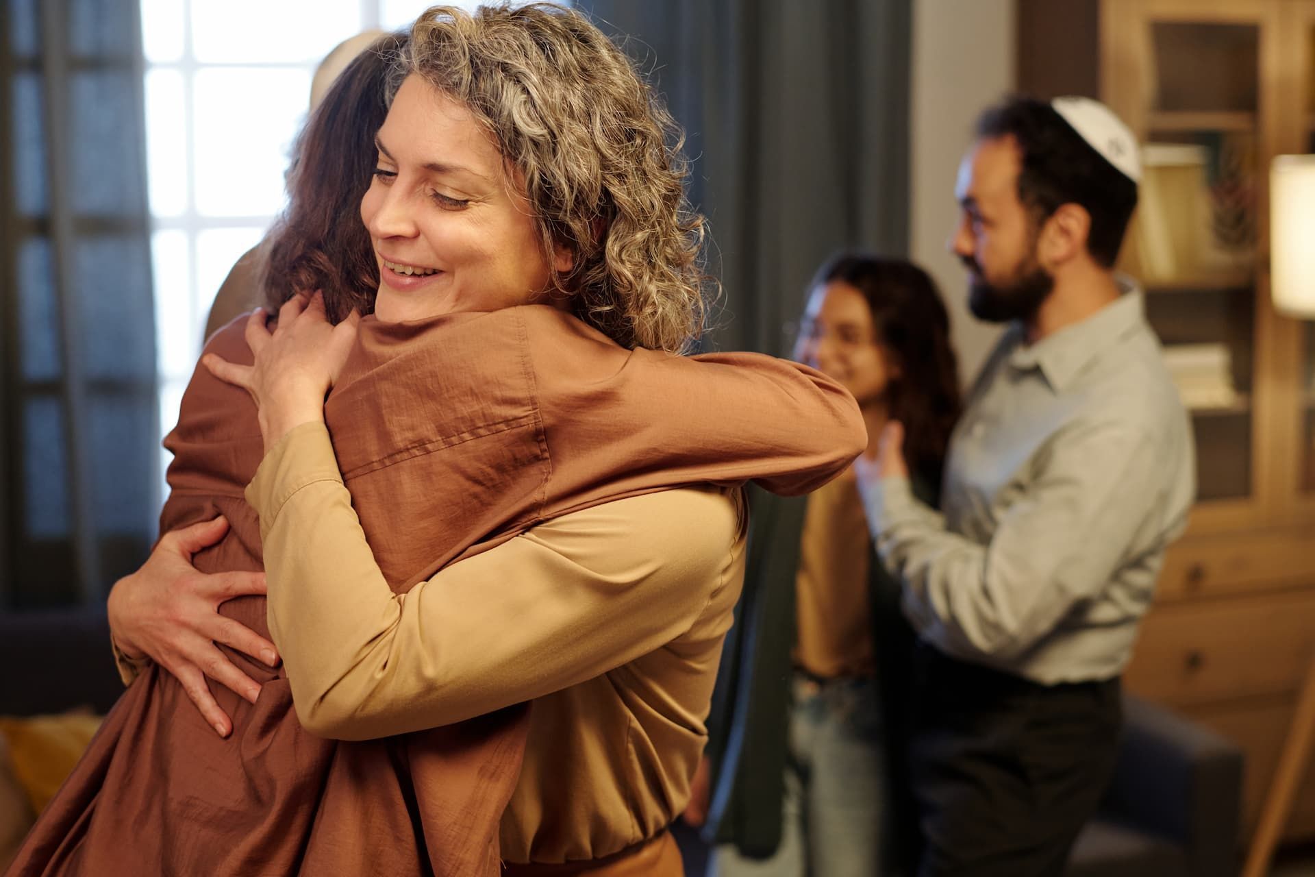 Two people hugging warmly in a cozy indoor gathering, with others chatting in the background.