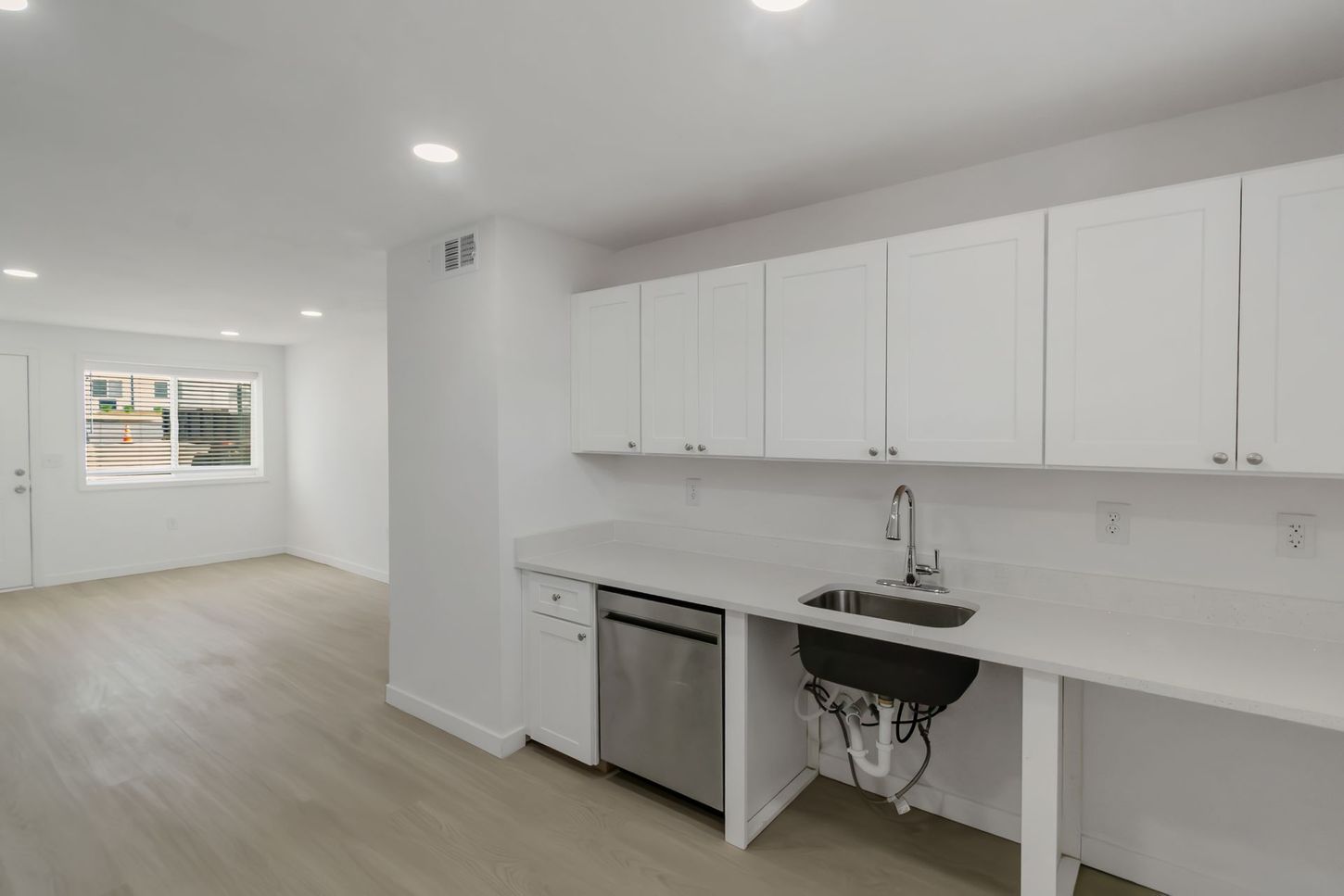 Bright modern kitchen with white cabinets, sink, dishwasher, and open living area with window.