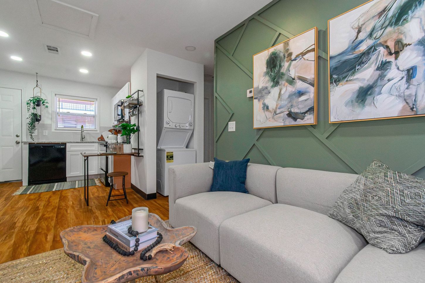 Modern living room with gray sofa, green accent wall, abstract art, and open kitchen beyond