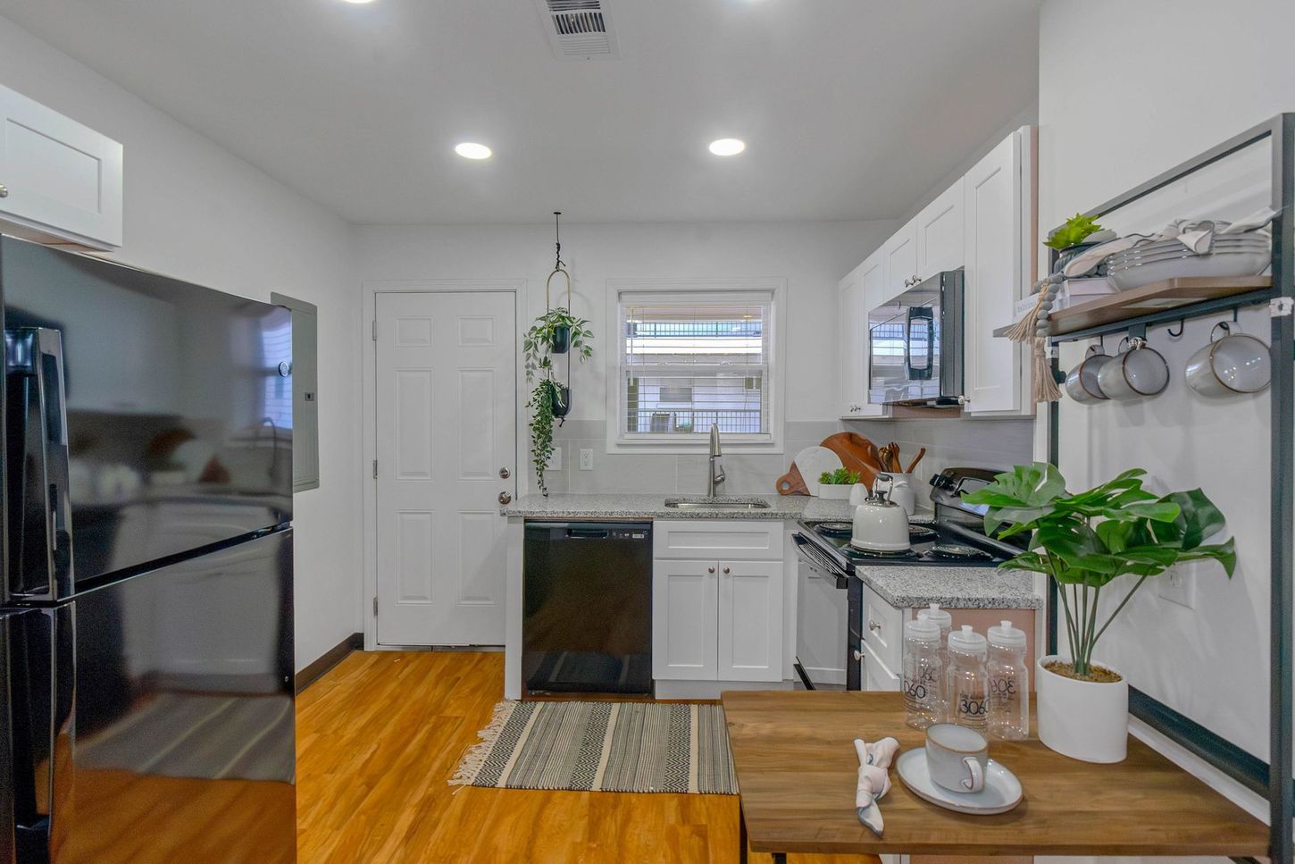 Bright white kitchen with black appliances, wood floor, and a small plant-filled counter by the sink.