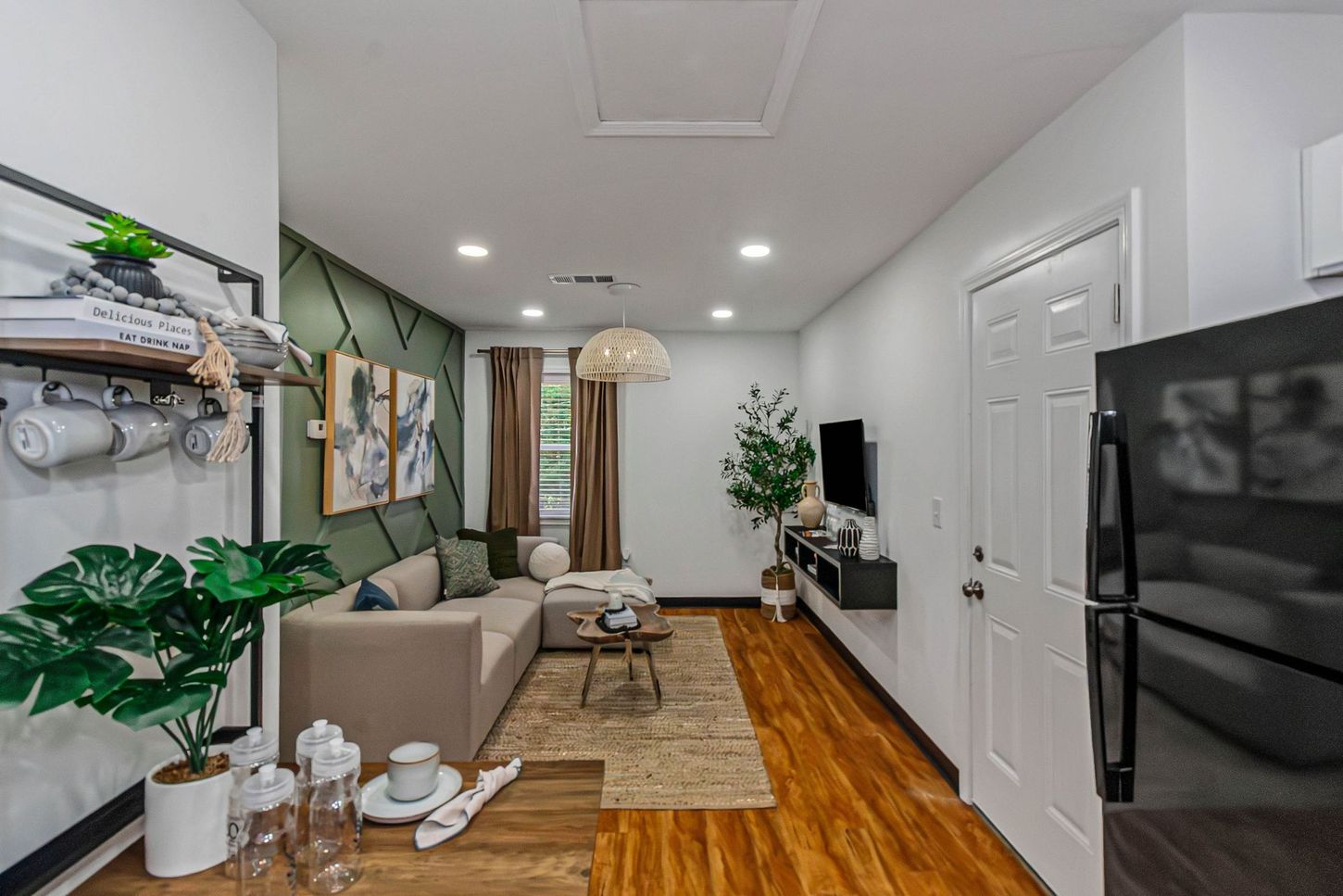 Bright living room with gray sofa, green plants, wood floor, and white kitchen cabinets