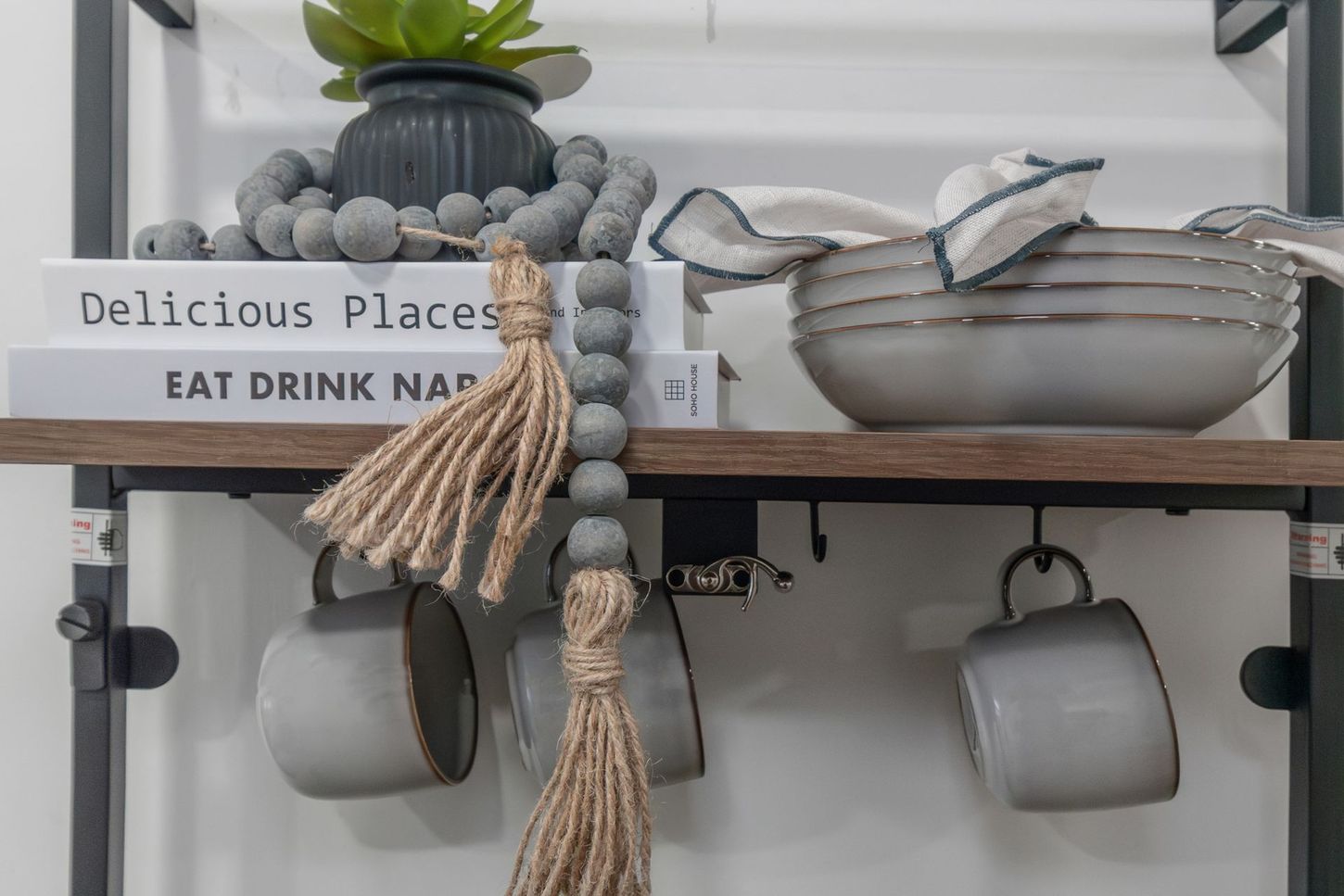 Decorative shelf with stacked books, hanging mugs, a potted plant, and a woven basket in a kitchen or café.