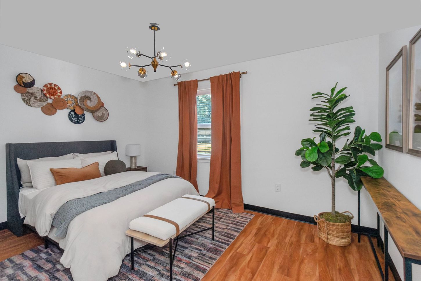 Bright bedroom with bed, bench, orange curtains, wood floors, and a large potted plant.