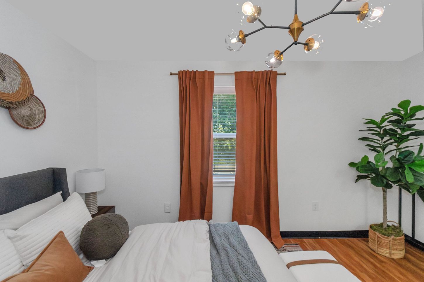 Bright bedroom with white bedding, orange curtains, wood floors, and a potted plant by the window