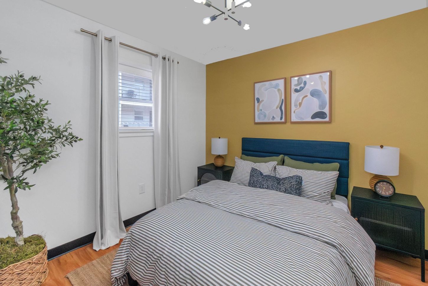 Bedroom with yellow accent wall, teal bed, striped bedding, and white curtains by a window.