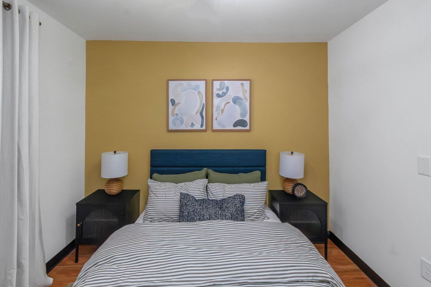 Bedroom with a bed, mustard accent wall, framed art, and matching bedside tables with lamps.