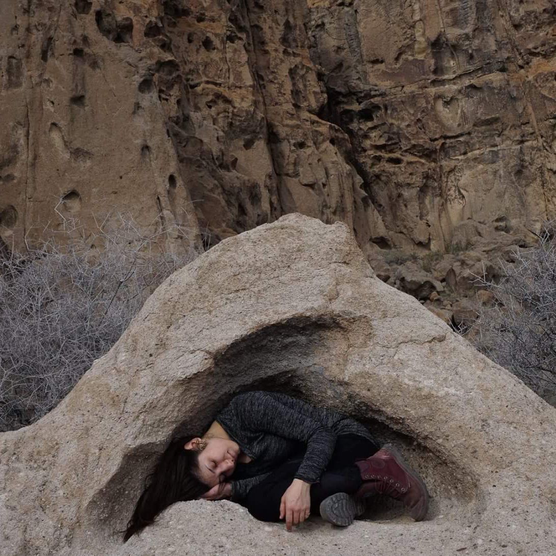 Person curled up sleeping inside a small rock cave beneath a cliff face.