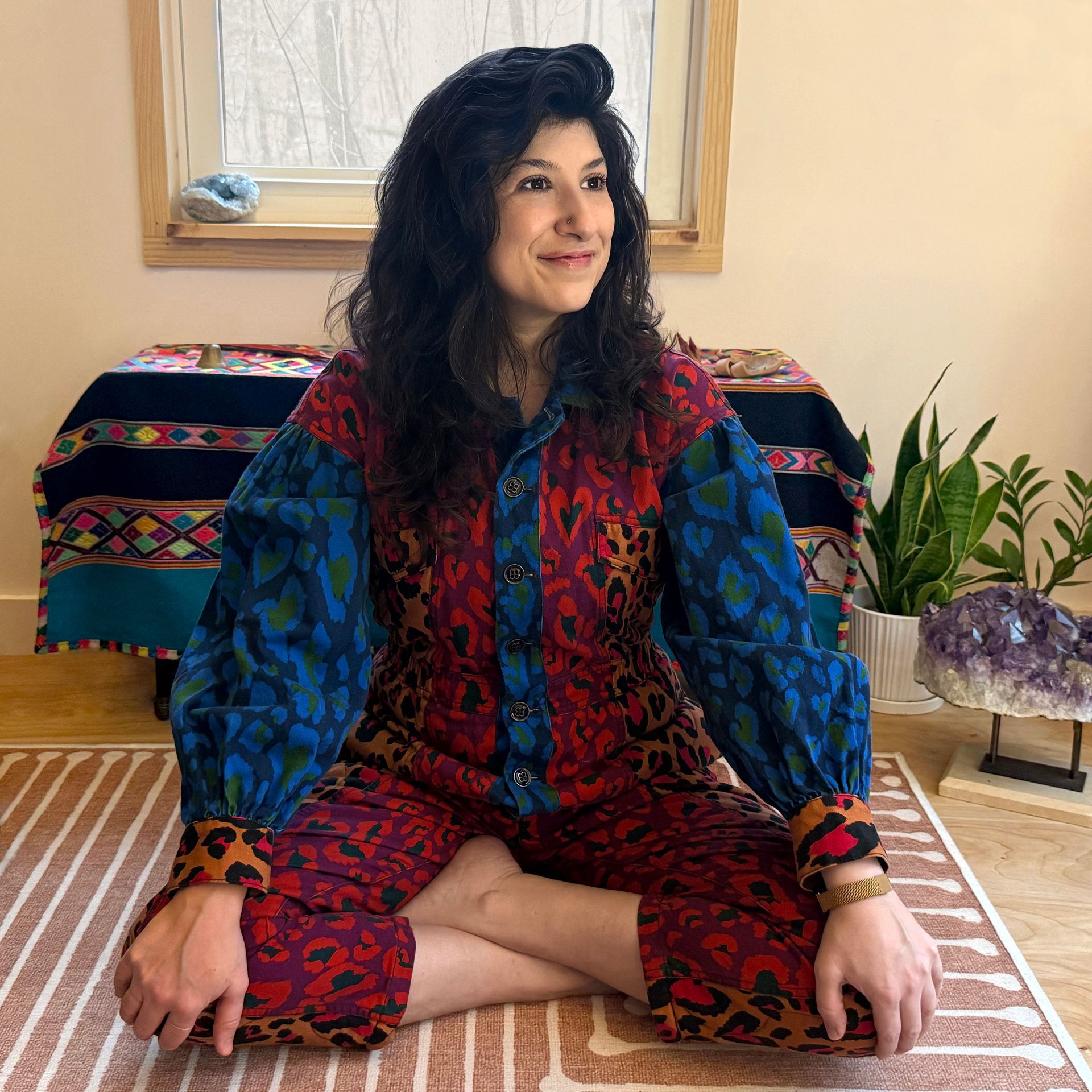 Woman seated cross-legged on a striped rug, wearing a colorful patterned outfit, in a bright room with plants.