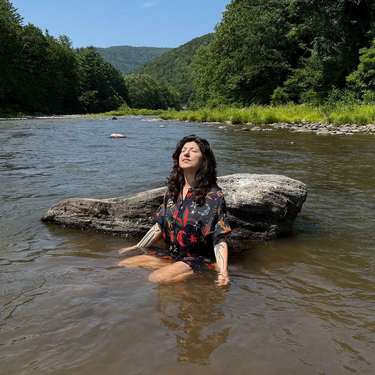 Picture of Cristina sitting in water in a blue tunic with their eyes closed and head tipped to the sun.
