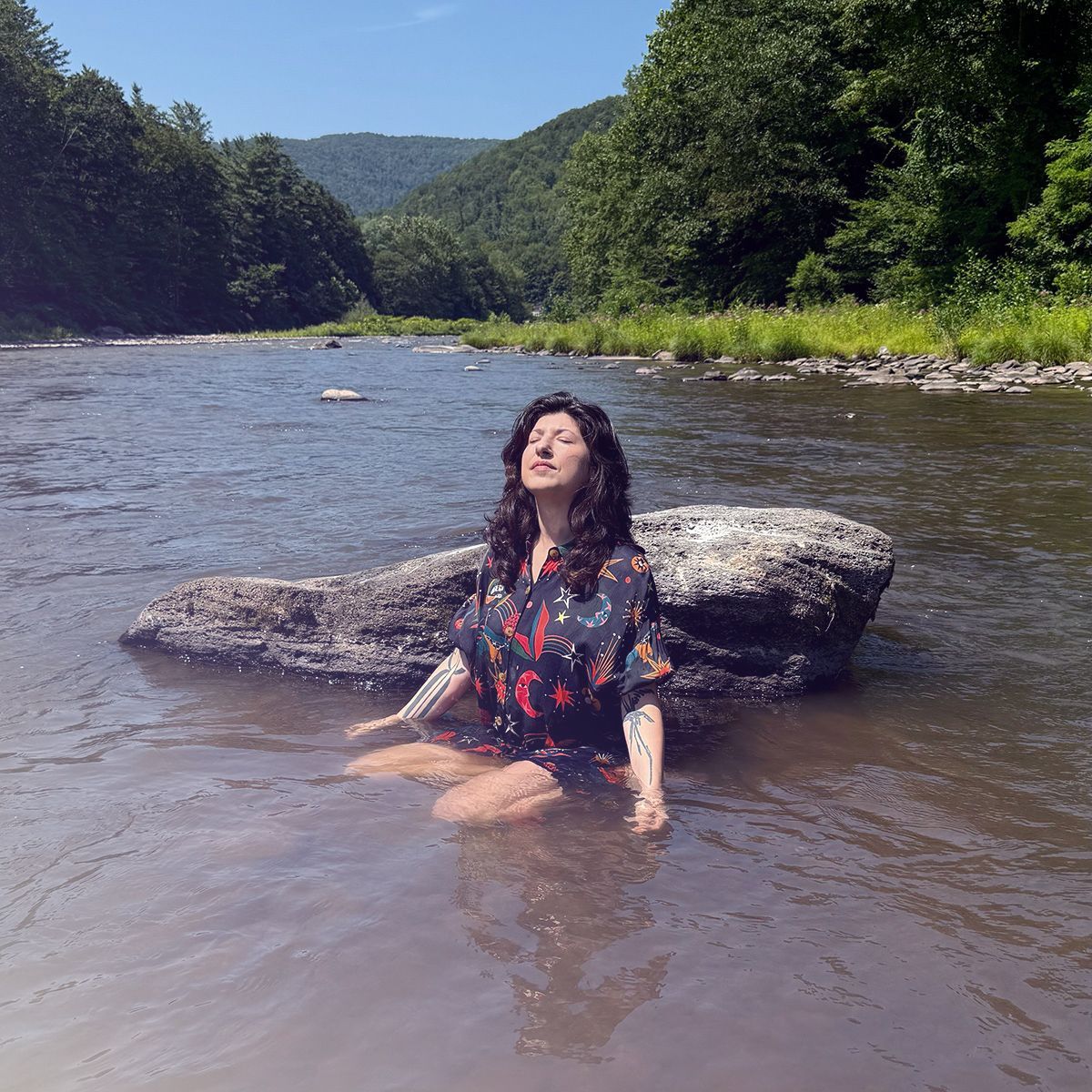Image of Cris sitting in the water against a rock with their eyes closed and head tilted up in the sun. 