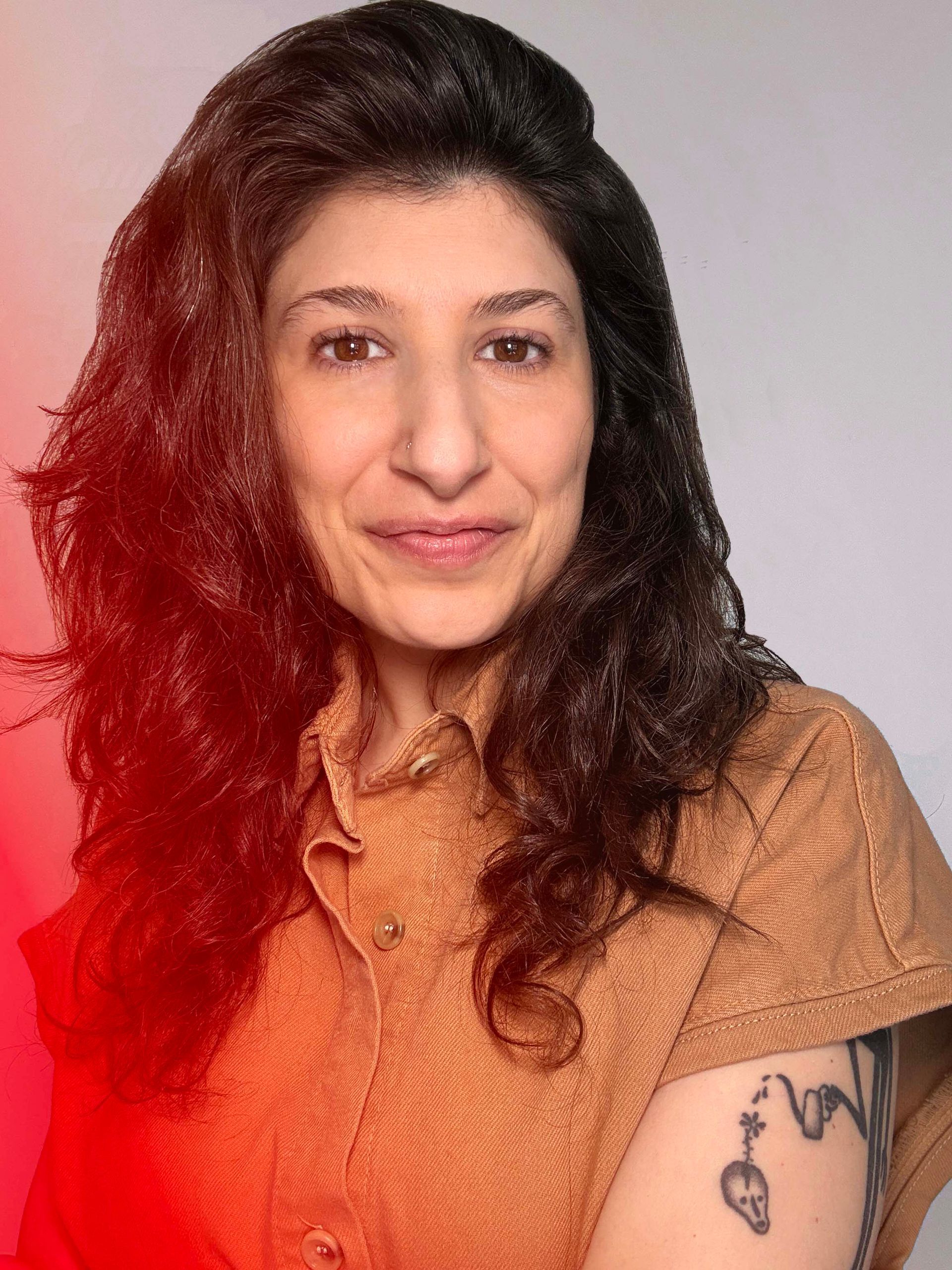 Portrait of a smiling person with dark curly hair, wearing a tan top, against a light background.