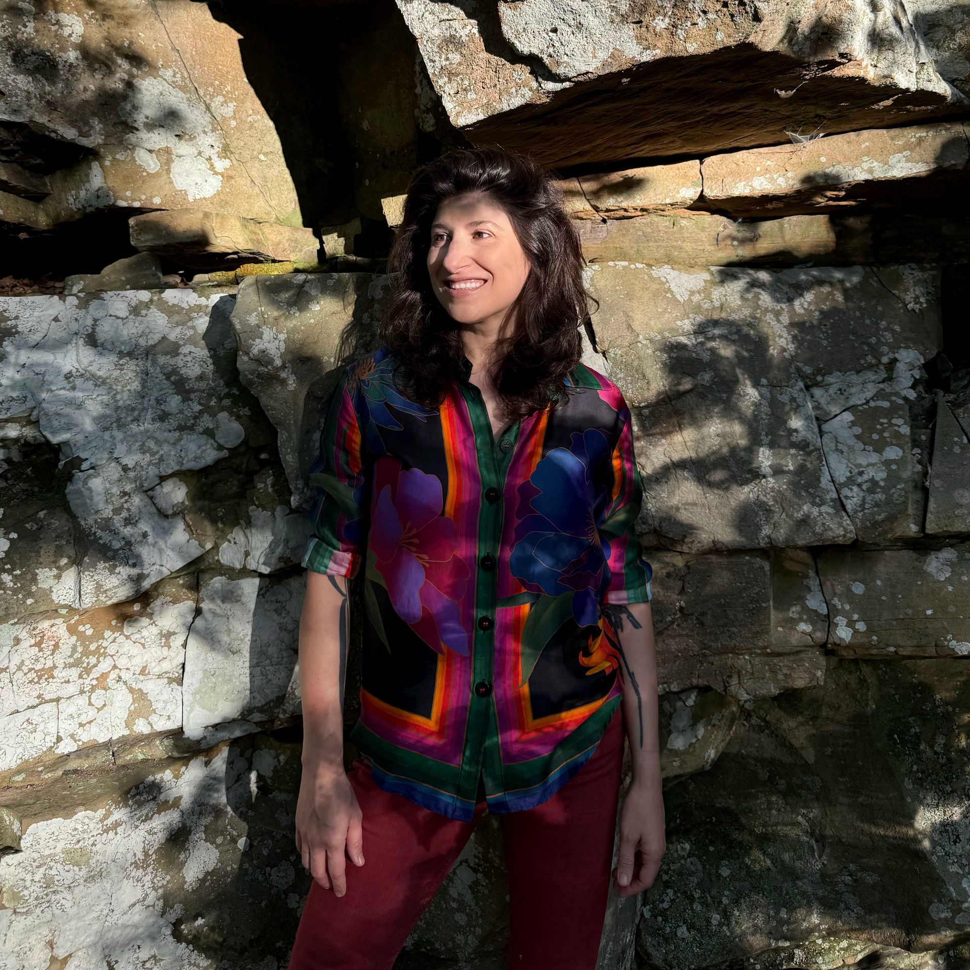 Woman in a colorful jacket standing by a rocky wall, smiling in sunlight.