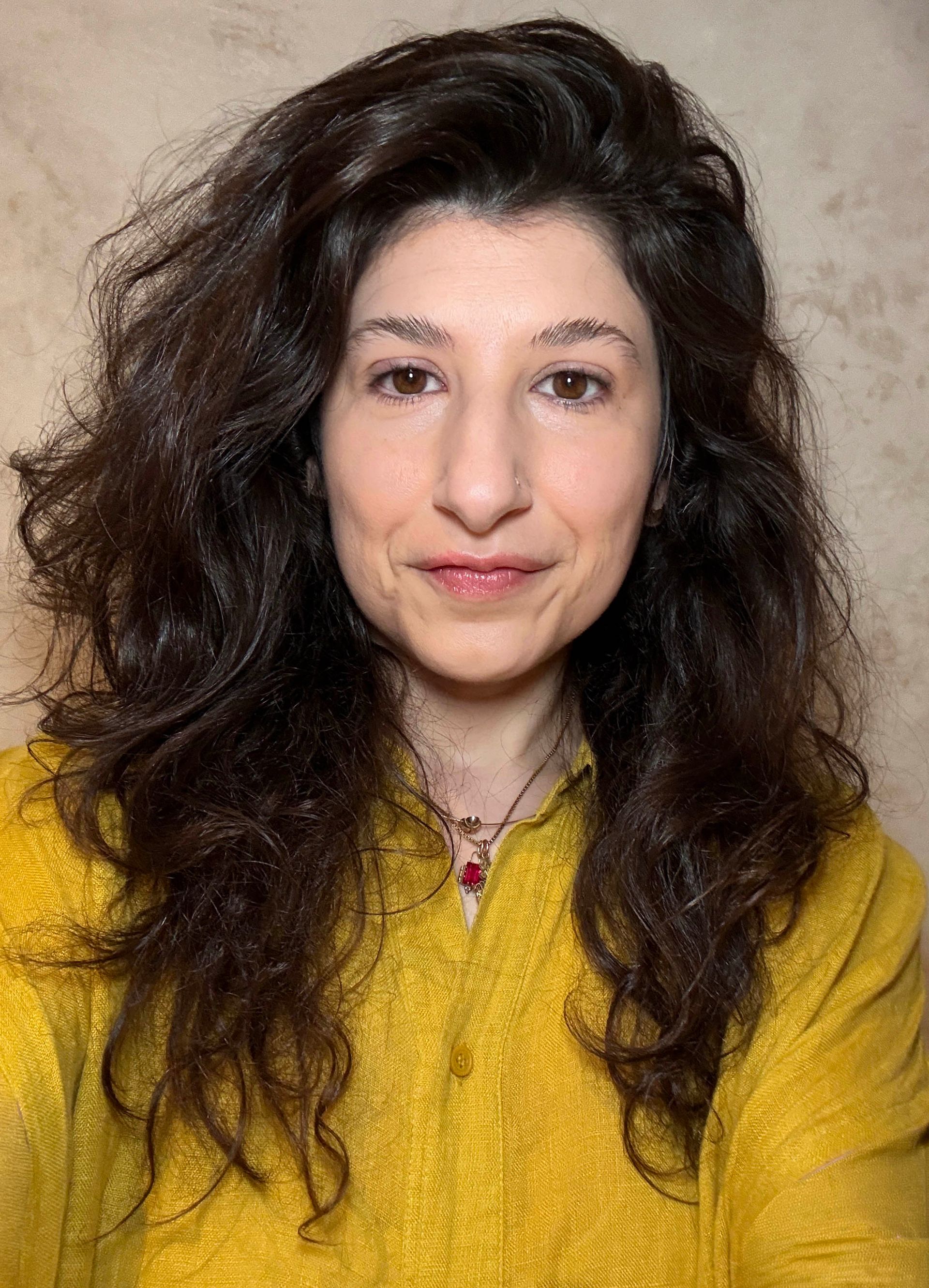 Woman with curly dark hair wearing a mustard yellow top, facing forward against a plain background