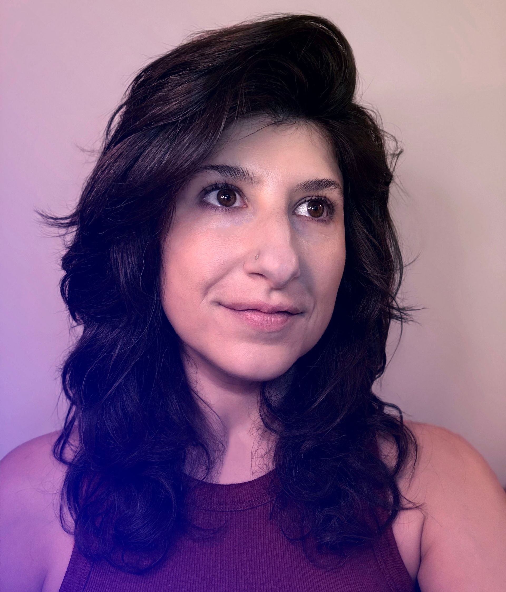 Portrait of a person with dark wavy hair, wearing a purple top, against a pink background.