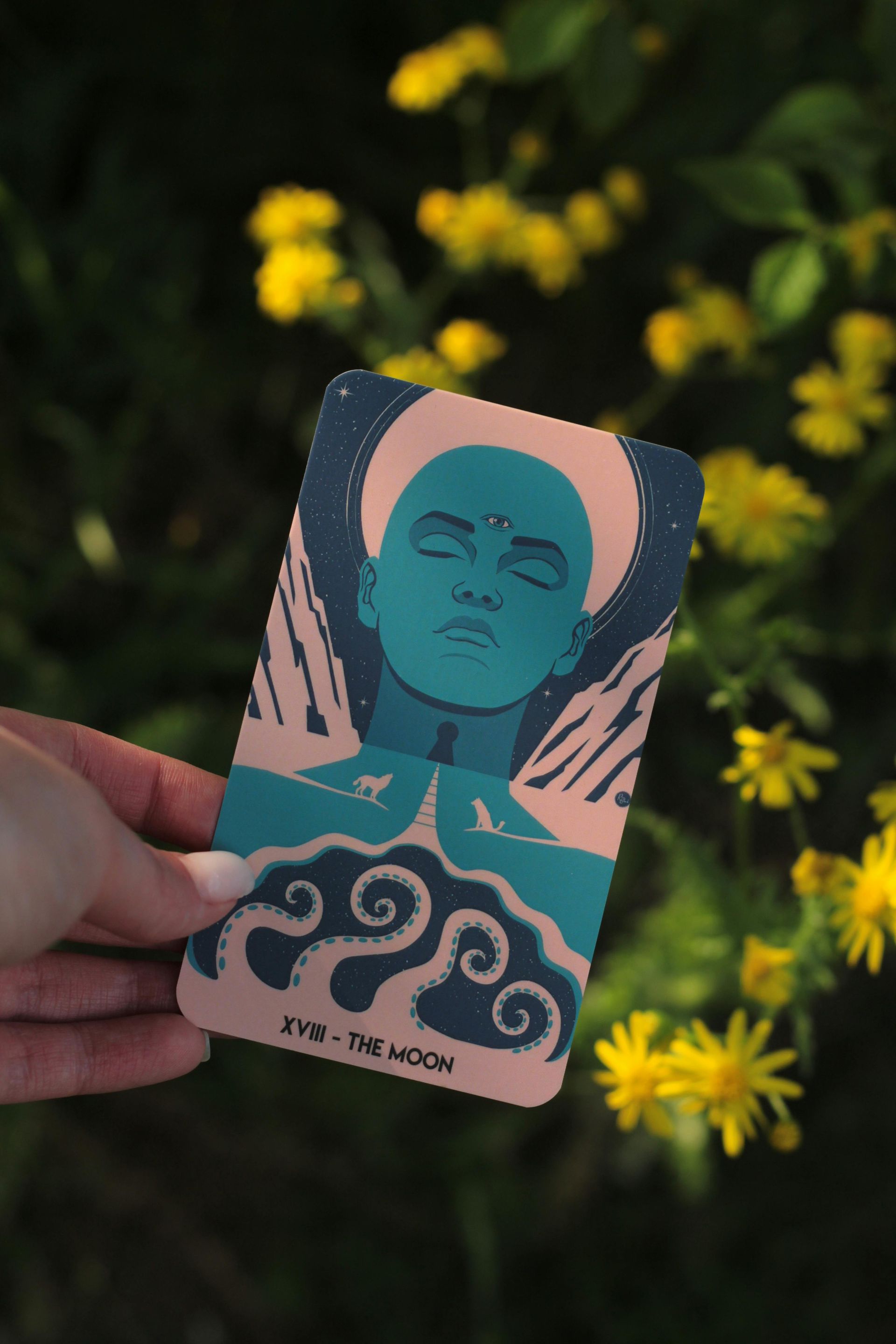 Hand holding a teal tarot-style card with a serene face above swirling waves, against yellow flowers.
