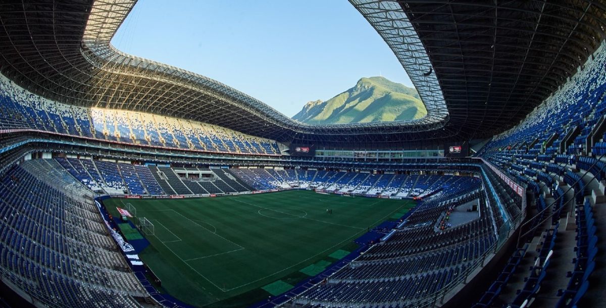 Vista panorámica de un estadio moderno con un campo de fútbol verde y un paisaje montañoso de fondo bajo un techo curvo.