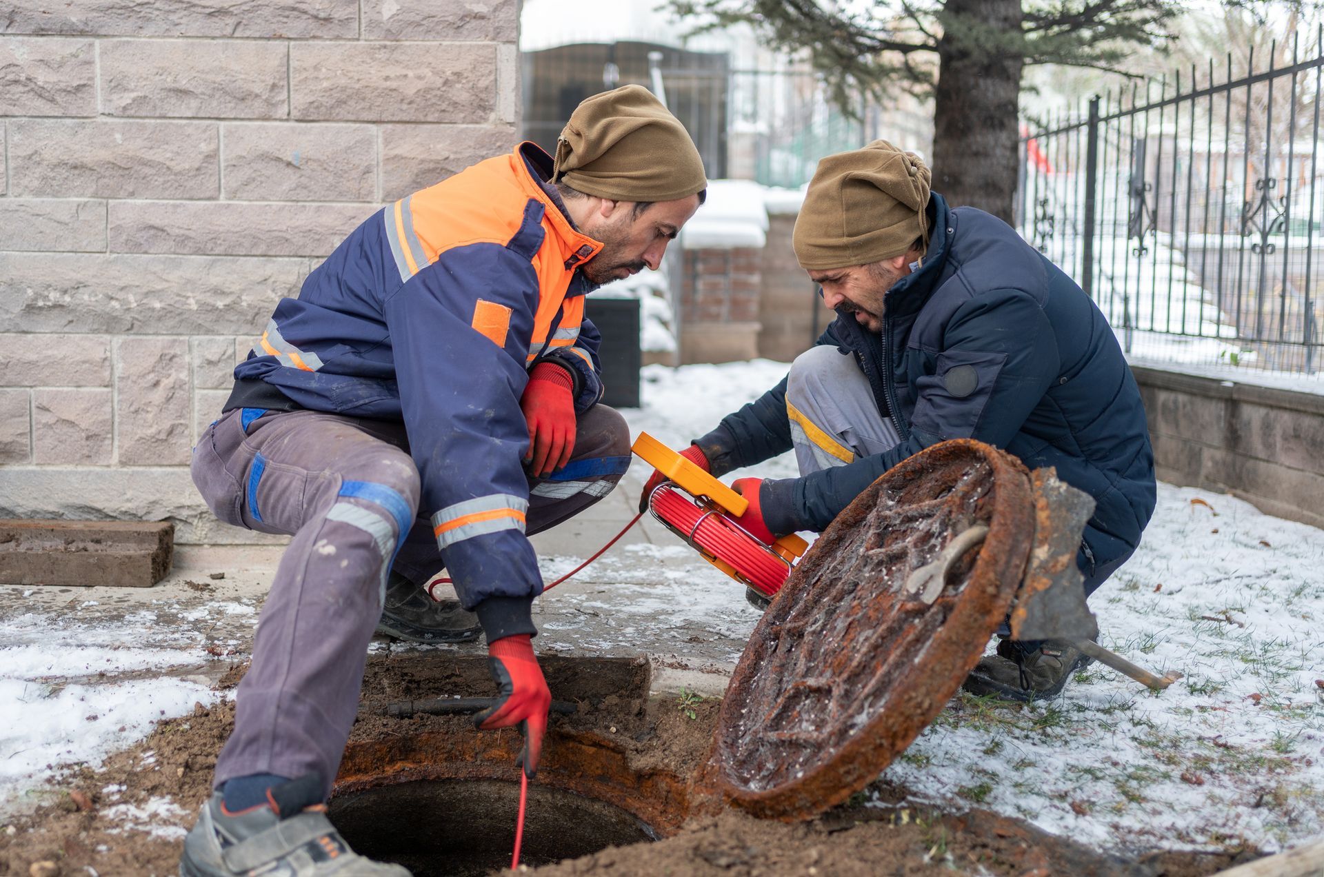 Two workers in safety gear examining a manhole, one using a device, outdoors near a brick wall.