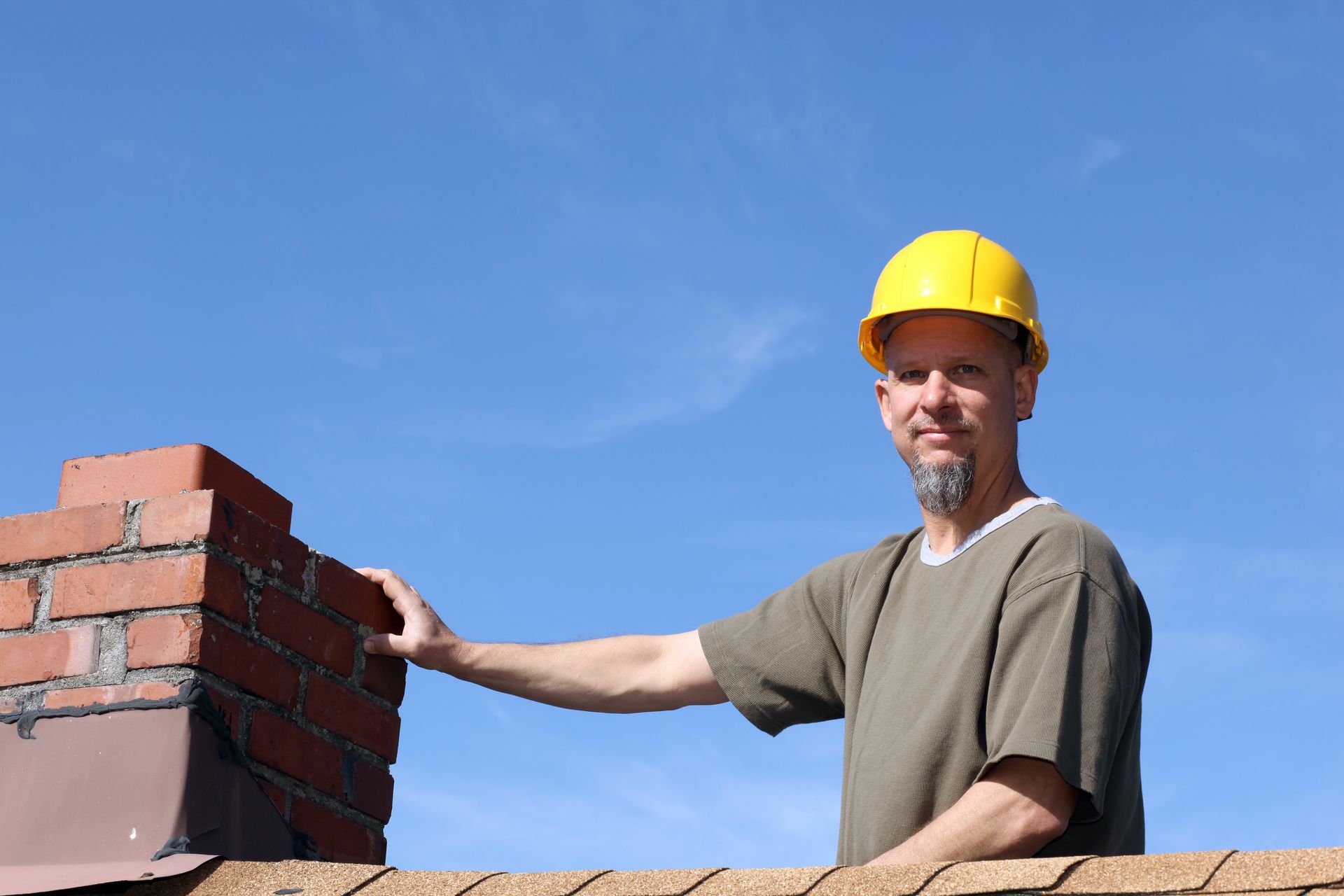 Man in yellow hard hat on a roof, next to brick chimney, blue sky background.