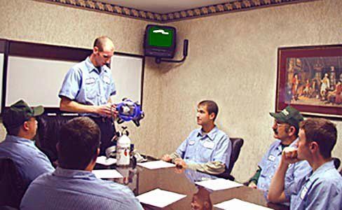 Group of men in blue shirts in a meeting, one demonstrating a machine, papers on the table.