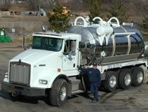 White septic tank truck with a man in blue standing beside it.