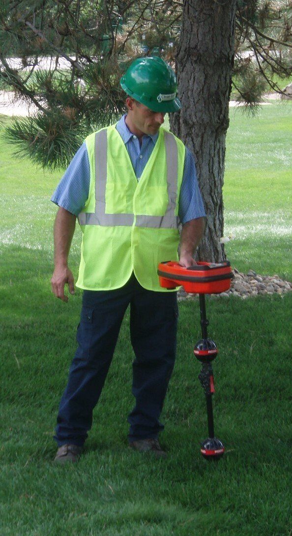 Septic Tank Locating – Kalamazoo, MI – Clean Earth Environmental Contracting Services Worker in safety vest uses a utility locator on grass near a tree.