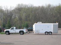 White pickup truck towing a white trailer on a paved area, trees in the background.